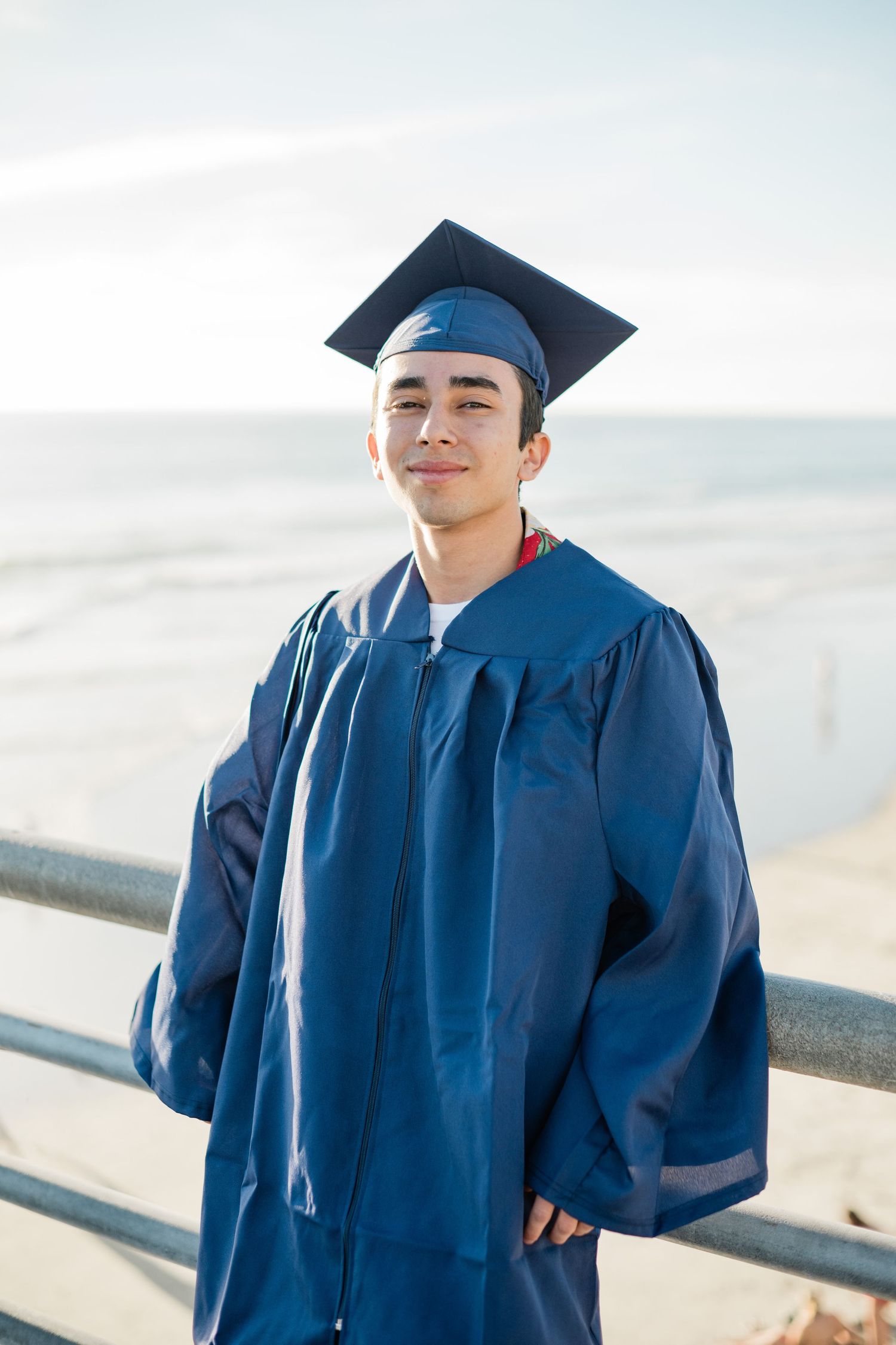 Young man in blue graduation cap and gown smiling outdoors near ocean railing on a sunny day.
