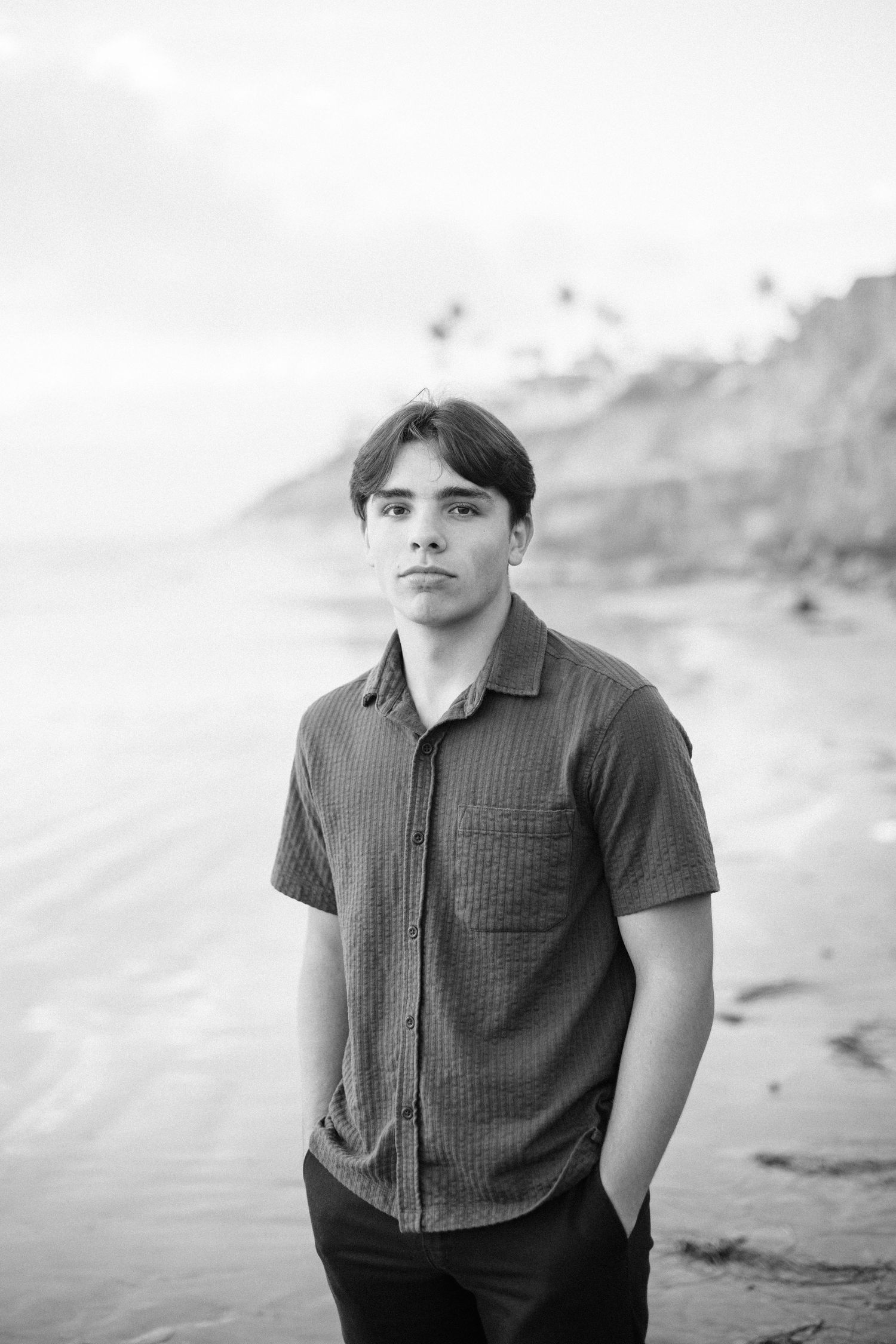 Black and white portrait of young man standing on beach in button-up shirt, cliffside background.