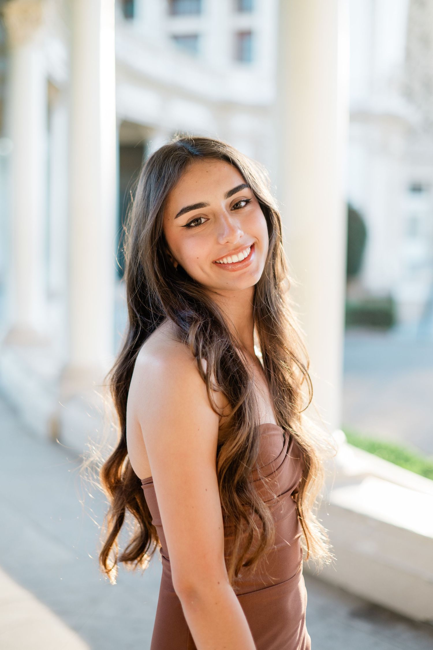 Young woman with long wavy brown hair smiling outdoors near white columns in a brown dress.