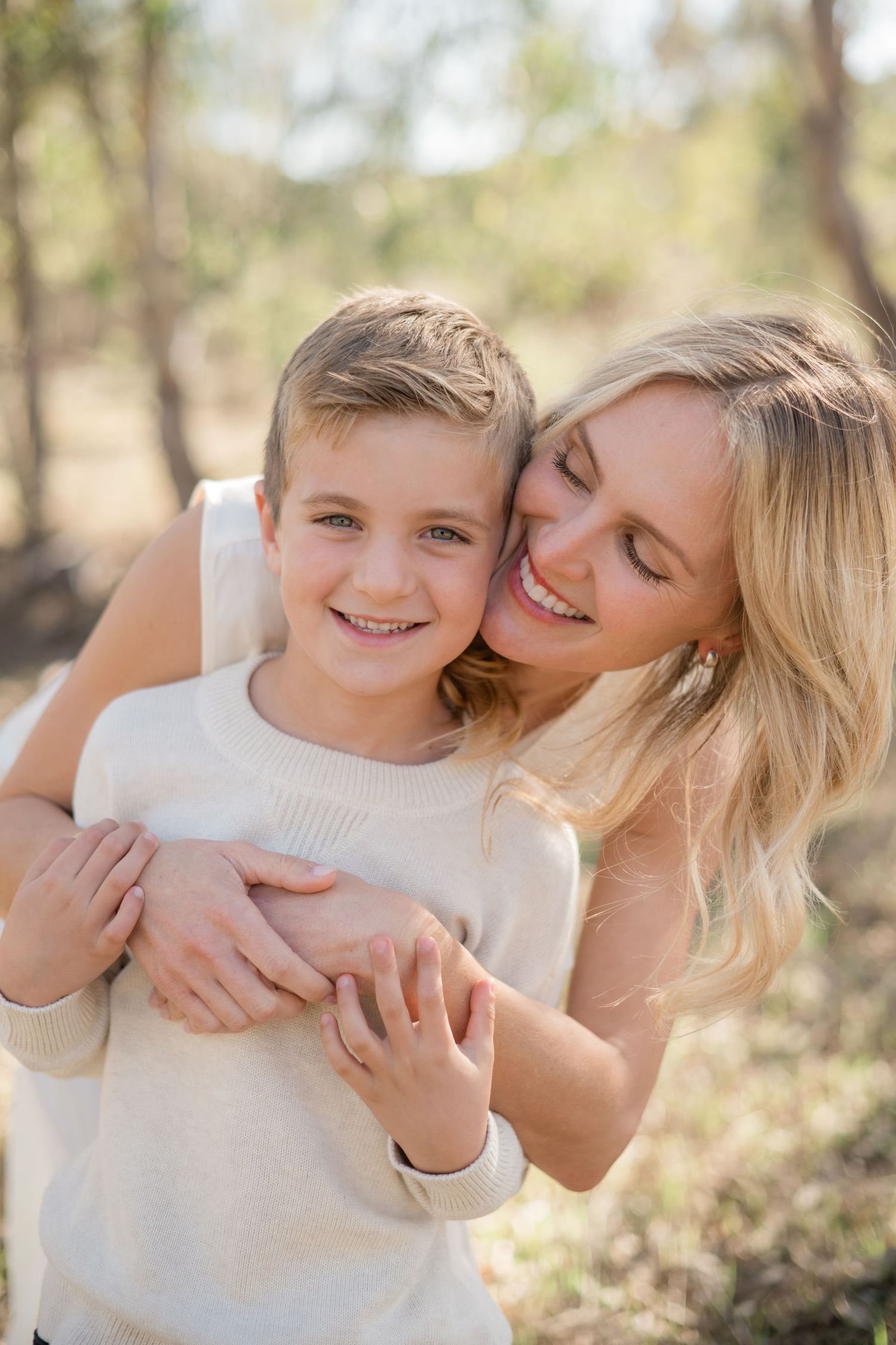 A smiling blonde woman hugging a happy young boy outdoors in a sunlit forest setting.