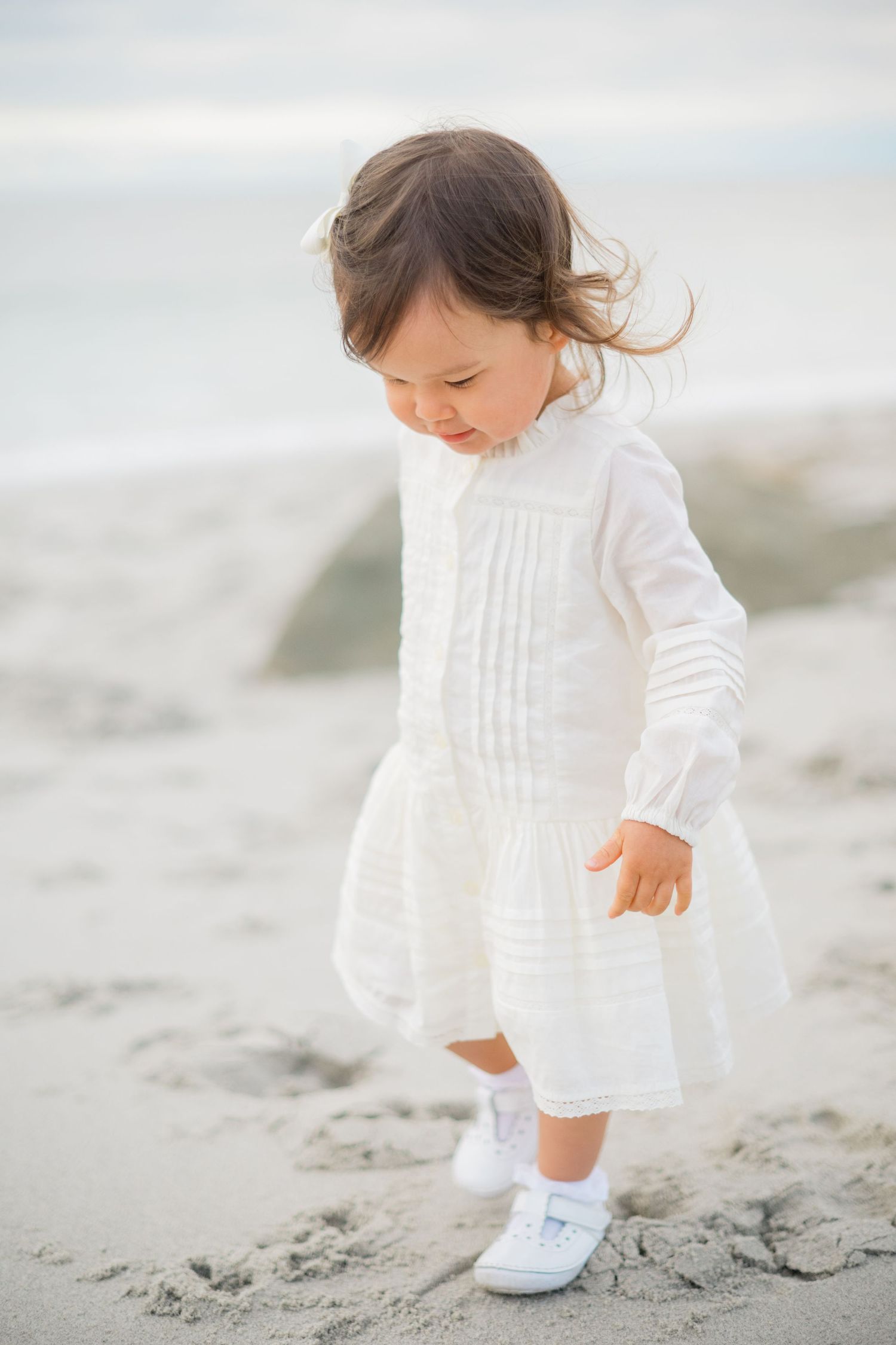 A cute toddler girl in a white dress explores the sandy beach shore.