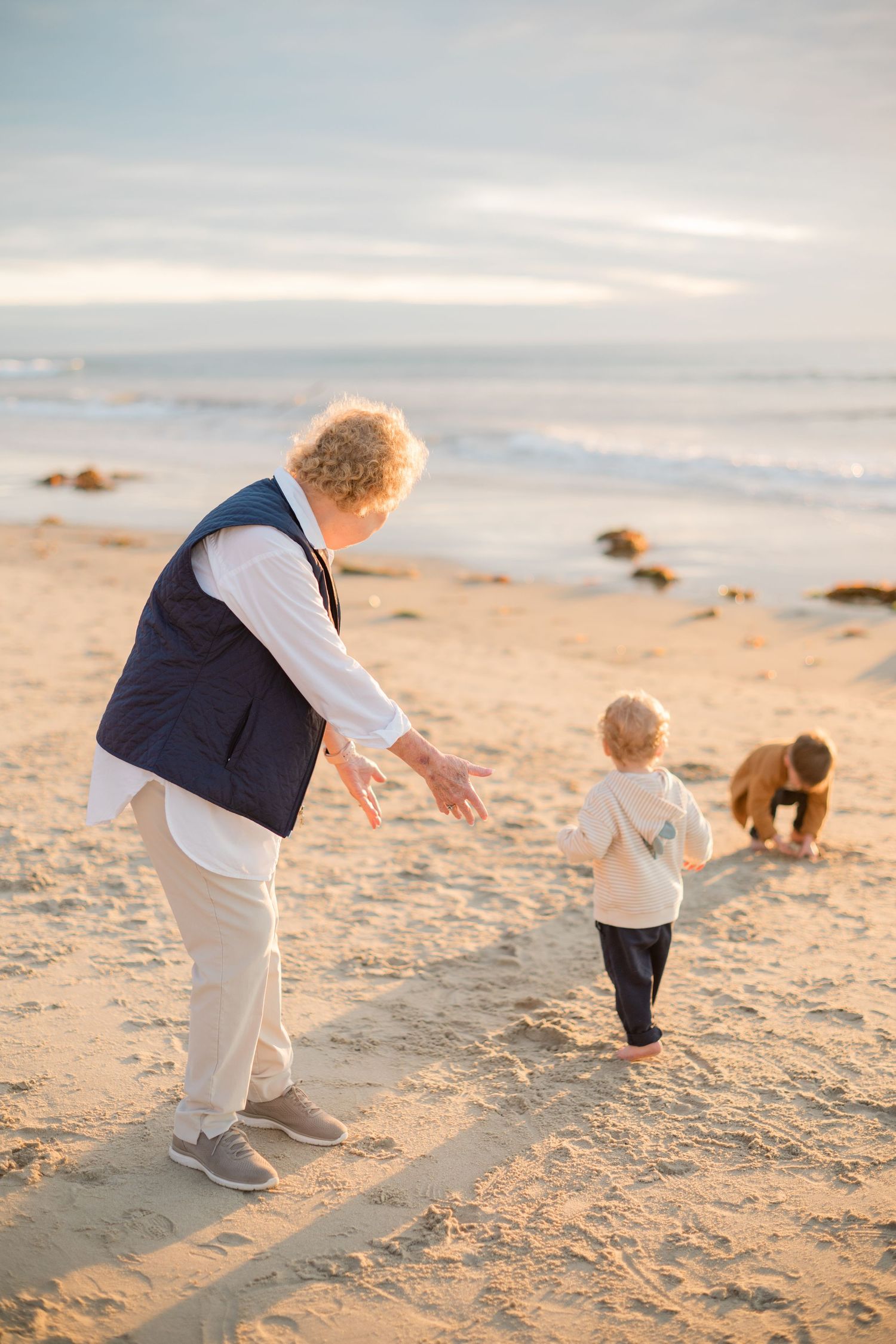 Grandmother reaching out to grandchildren playing on a sunny beach with ocean waves in the background.