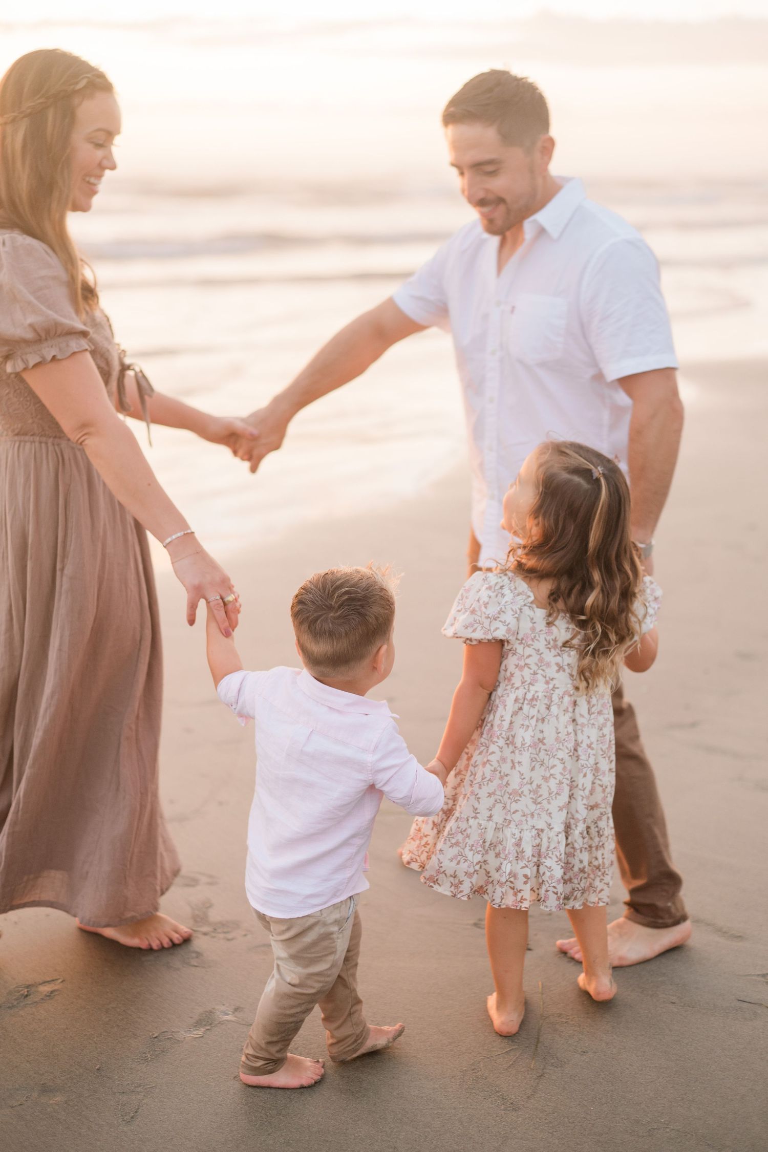 Family of four holding hands in a circle on a sandy beach at golden hour sunset.