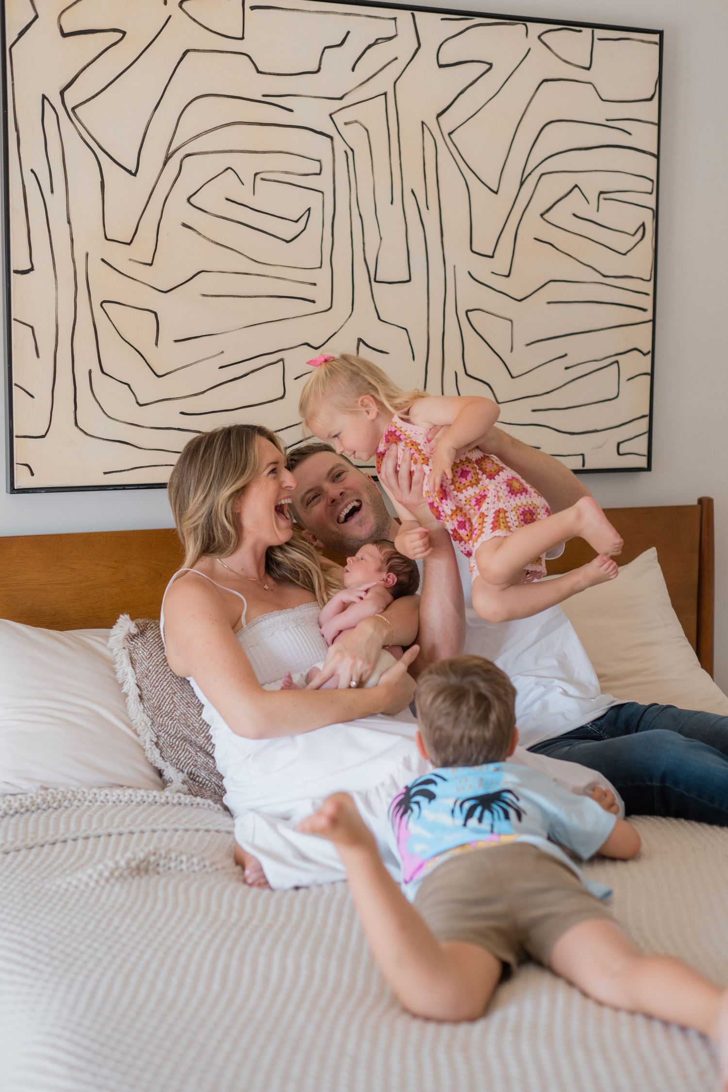 Happy family with newborn baby laughing together on bed, with abstract black and white wall art in background.