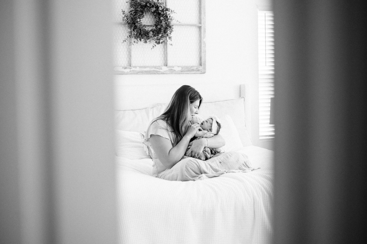 Black and white photo of a mother sitting on a white bed, tenderly holding her newborn baby in a bright room.
