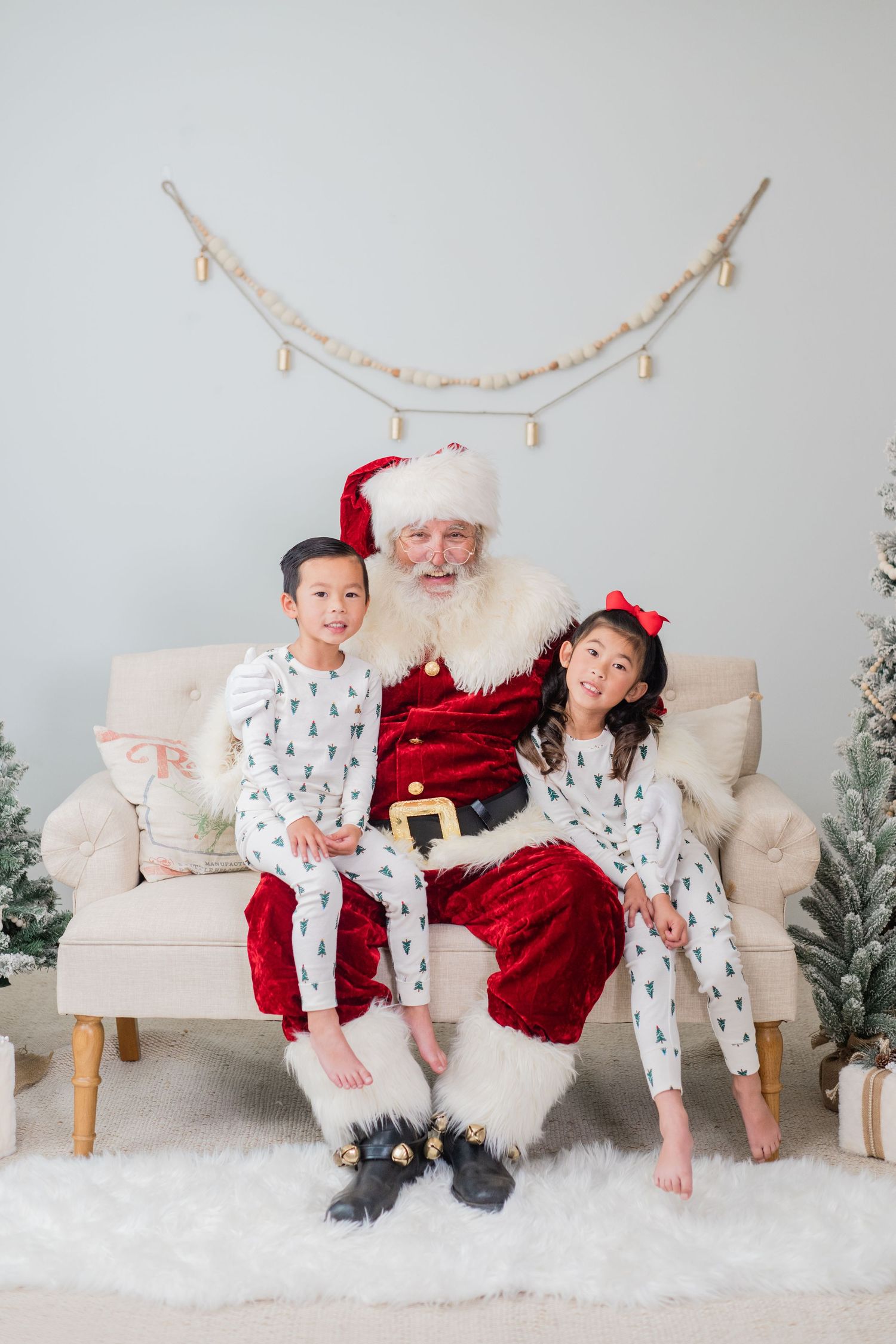 Two children in white Christmas tree pajamas sit on Santa Claus's lap on a cozy beige sofa.