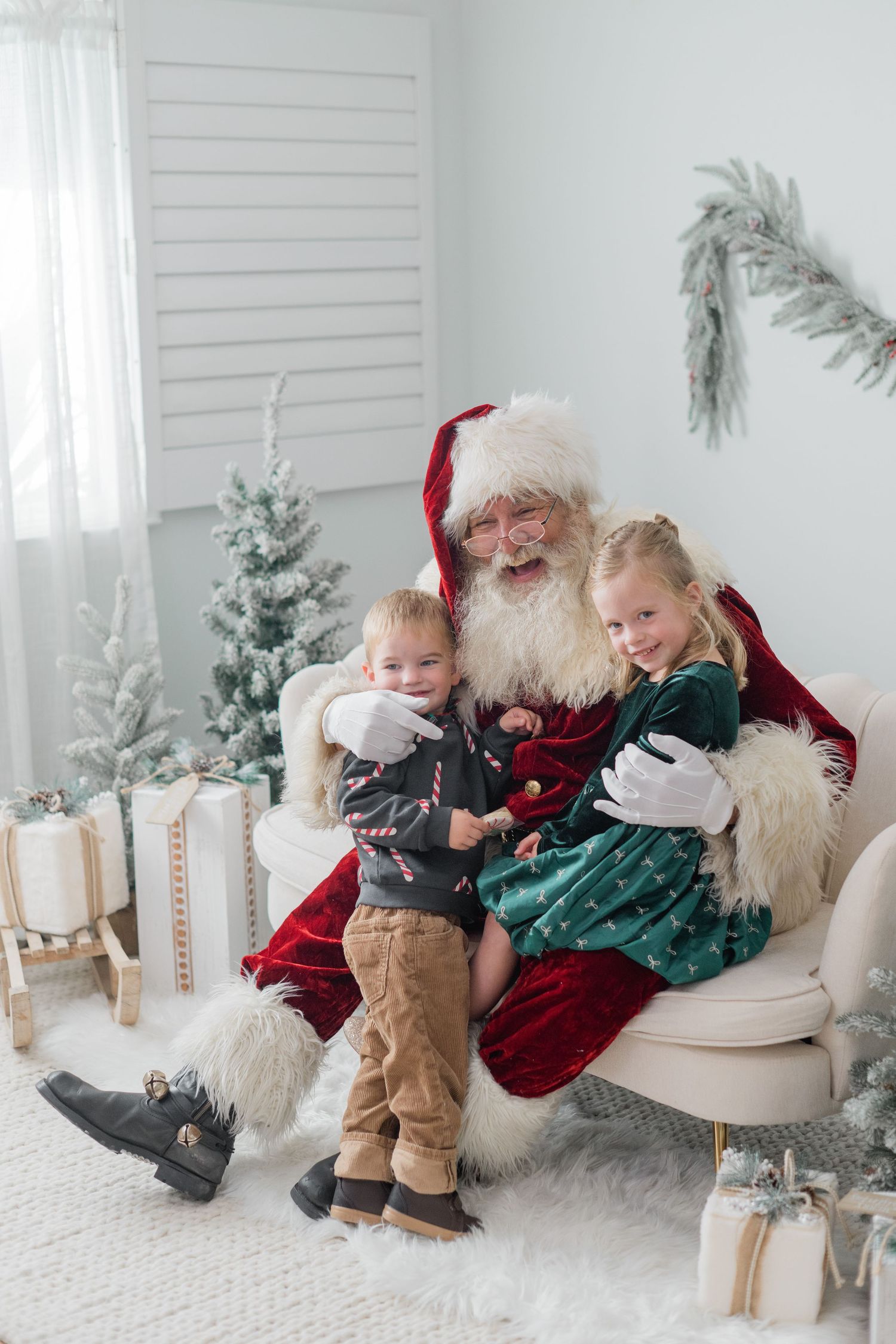 Santa Claus laughs while hugging two young children on a white armchair surrounded by Christmas gifts.
