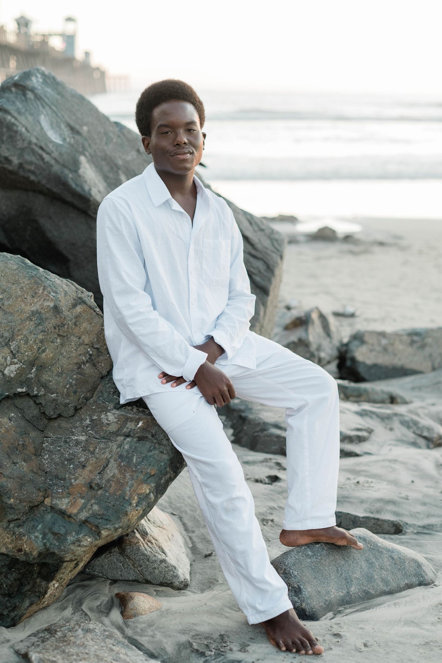 Young Black man in white linen outfit sitting barefoot on rocks at a sandy beach during golden hour.