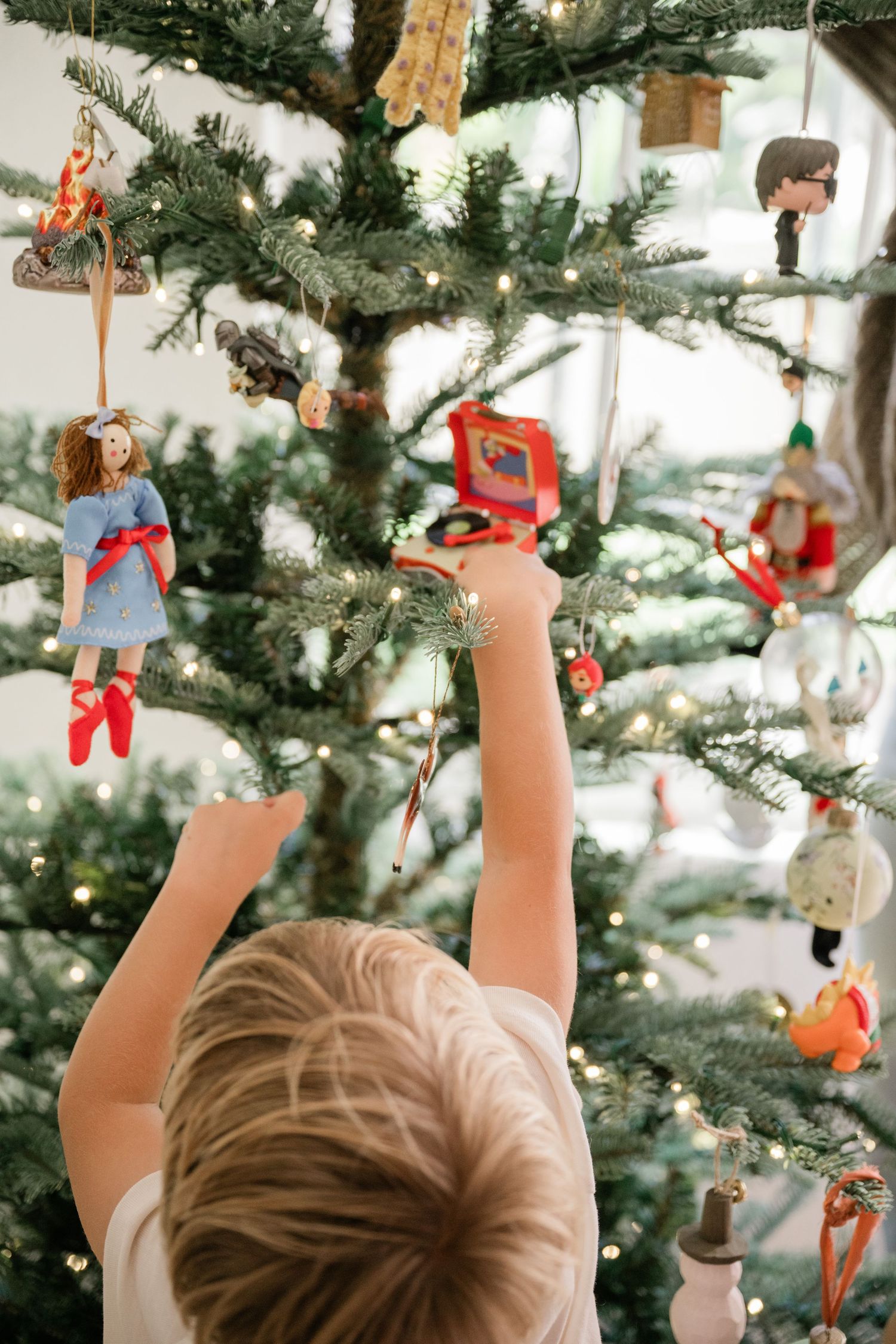 Young child reaching up to hang ornaments on a decorated Christmas tree with lights.