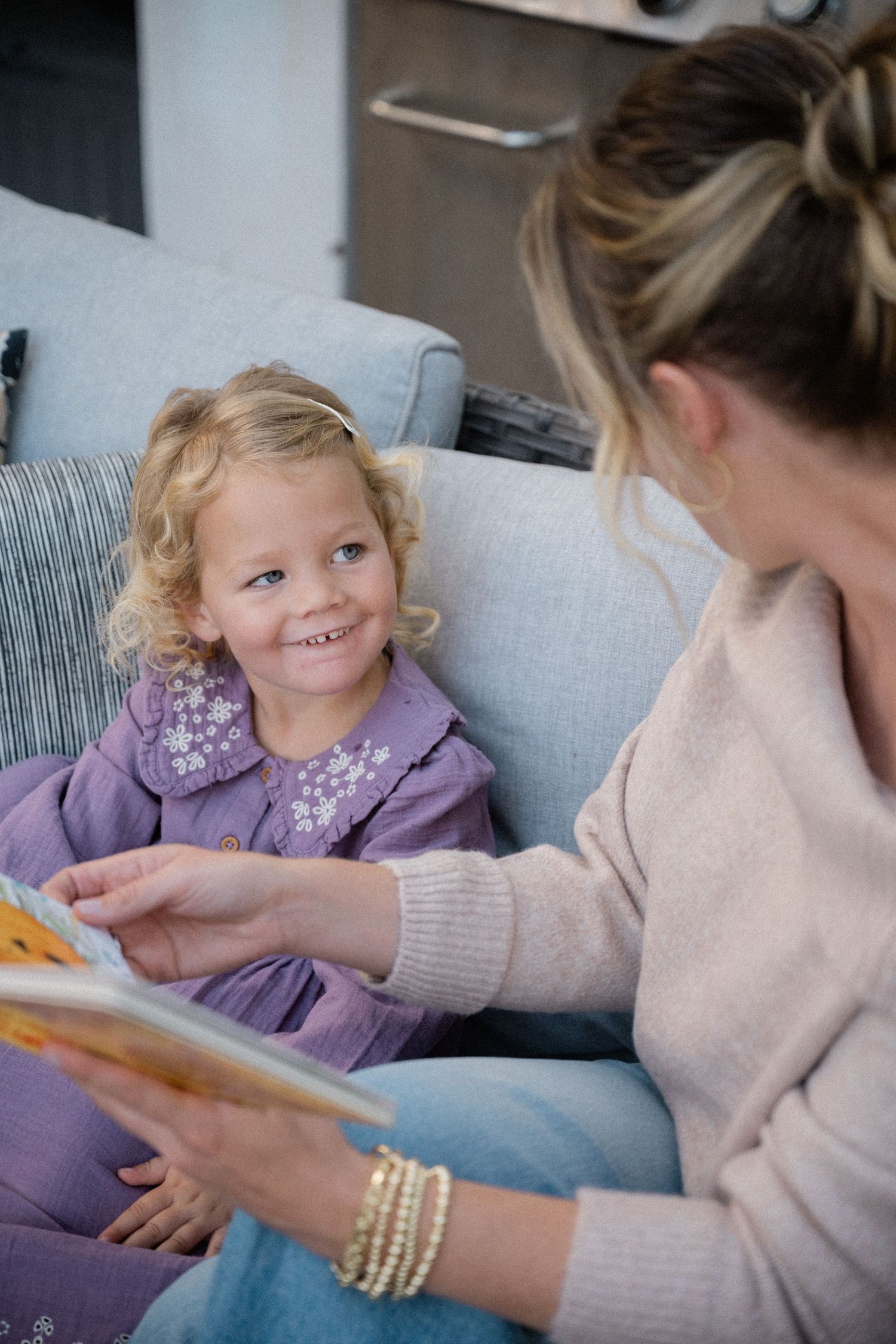 A smiling blonde toddler girl in purple dress sits on sofa while woman reads colorful picture book to her.