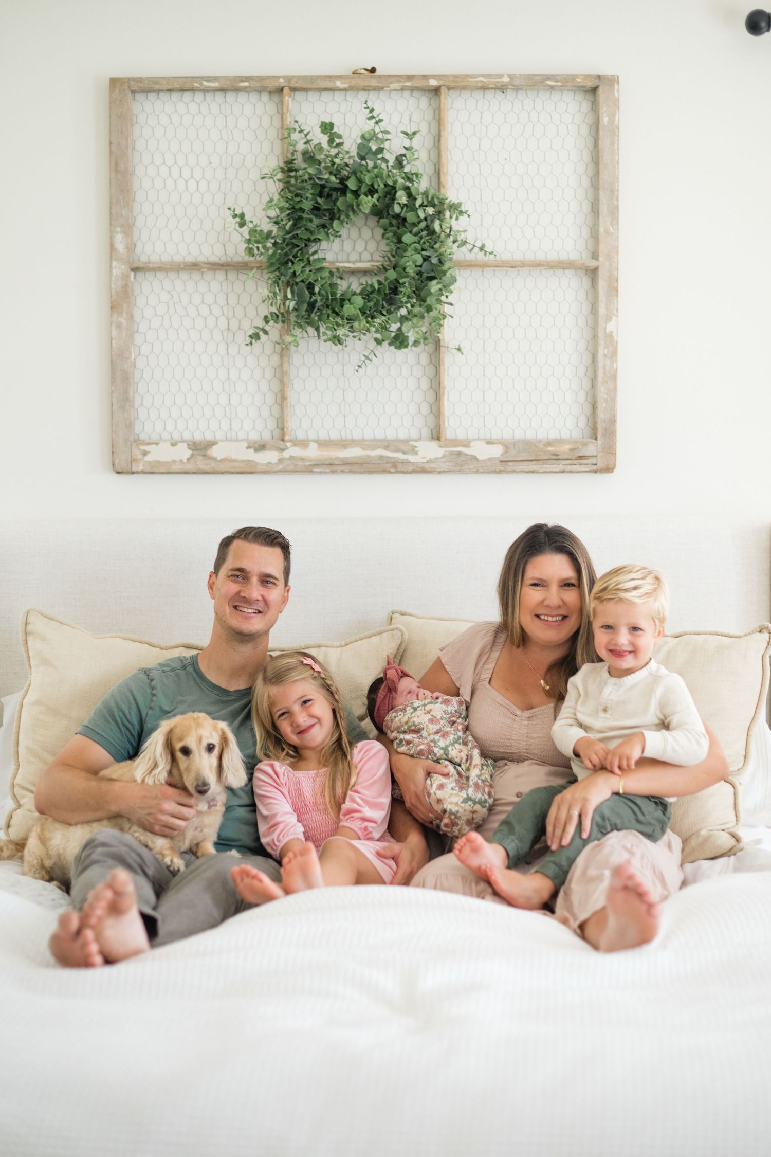 Happy family with three kids and a dachshund sitting on a white bed beneath a rustic window frame with a green wreath.