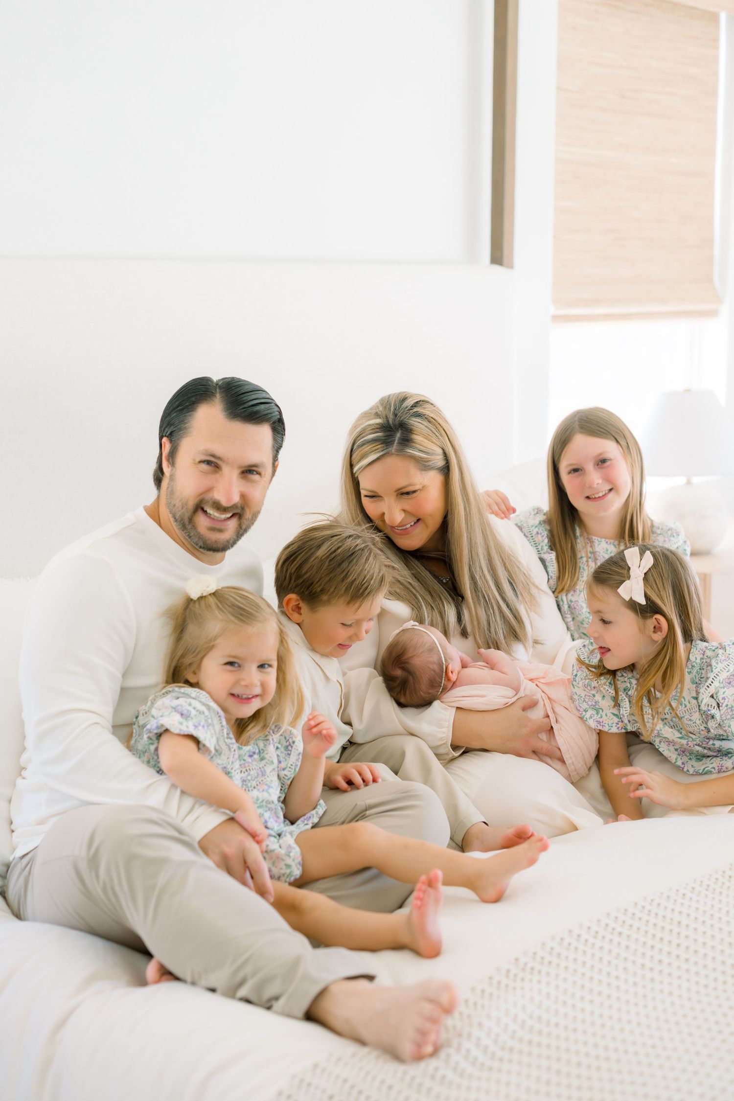 Happy family of seven with parents, four children, and a newborn baby posing together on a white bed.