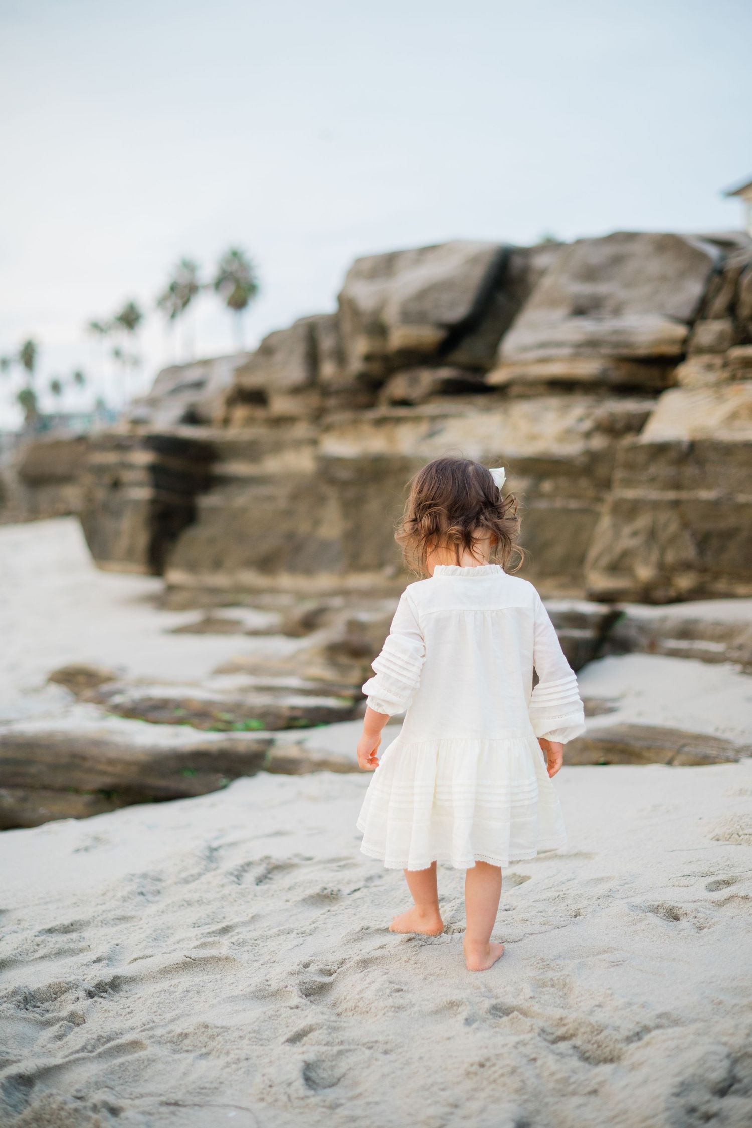 Young toddler girl in white dress walks barefoot toward rocky beach cliffs from behind.