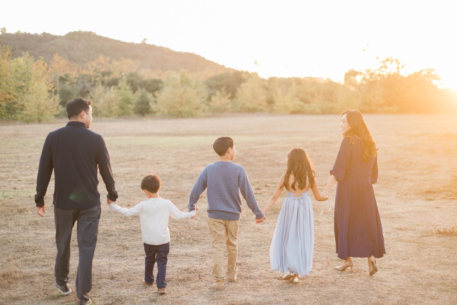 Family of five walking hand-in-hand through golden field at sunset with rolling hills in background.