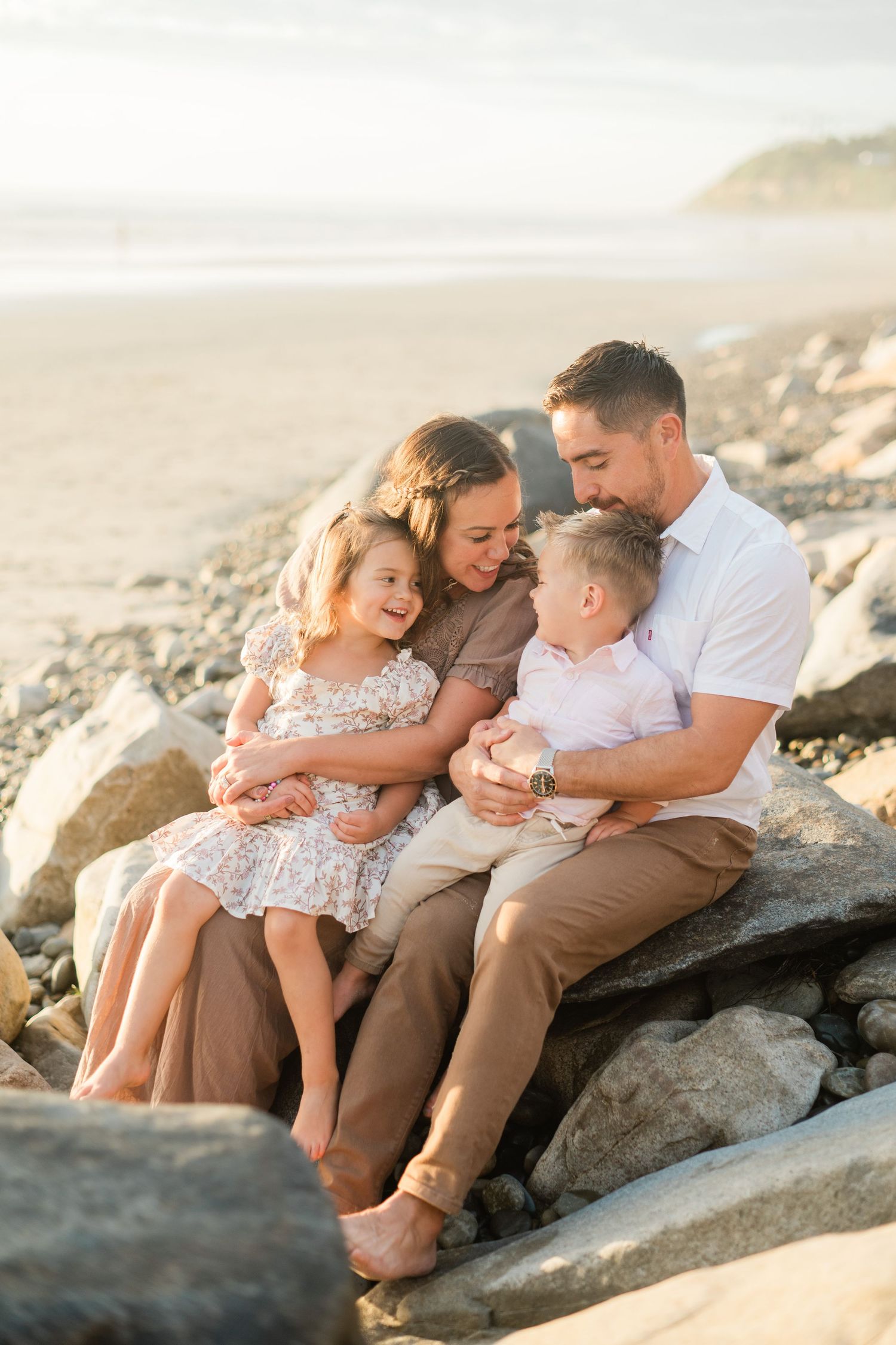 Family of four sitting on rocky beach at golden hour, parents embracing young daughter and toddler son warmly.