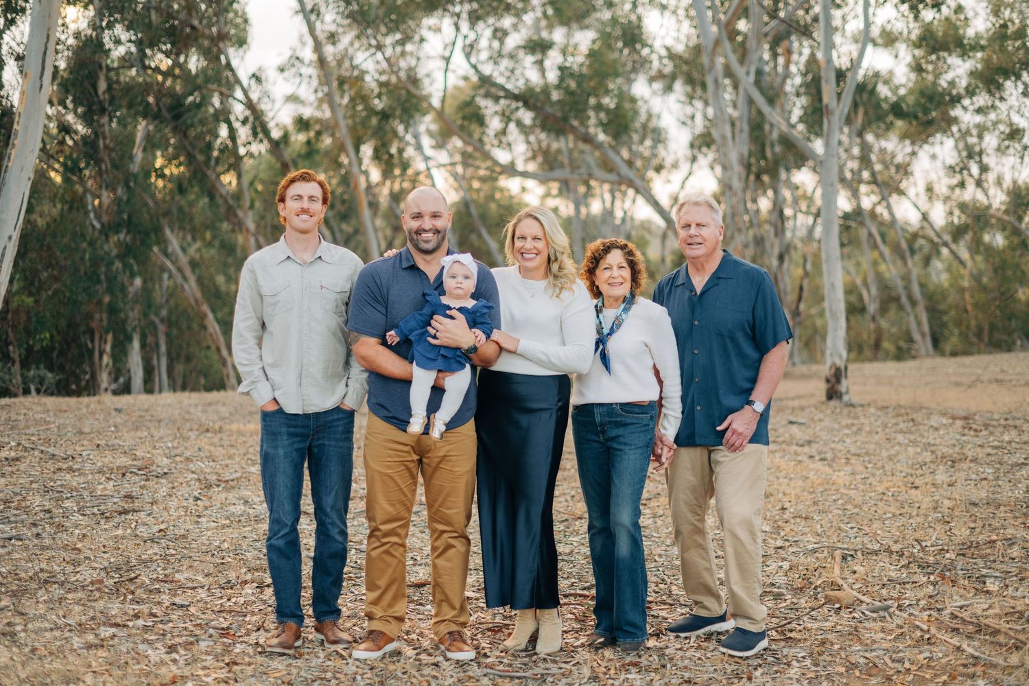 Family of six posing outdoors among eucalyptus trees, including a baby, smiling together on a sunny day.