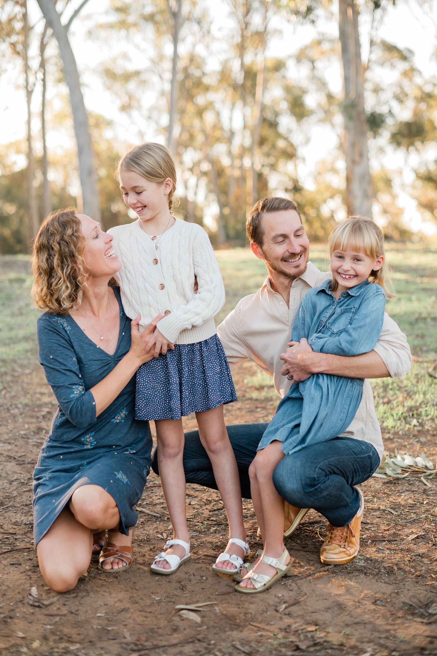 Happy family of four posing outdoors in a sunlit park, parents kneeling with two smiling blonde daughters.
