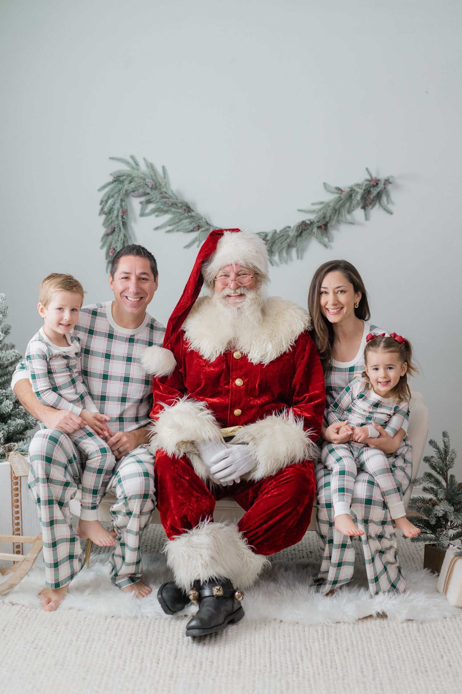 Family in matching plaid pajamas posing with Santa Claus in a festive Christmas photo studio setting.