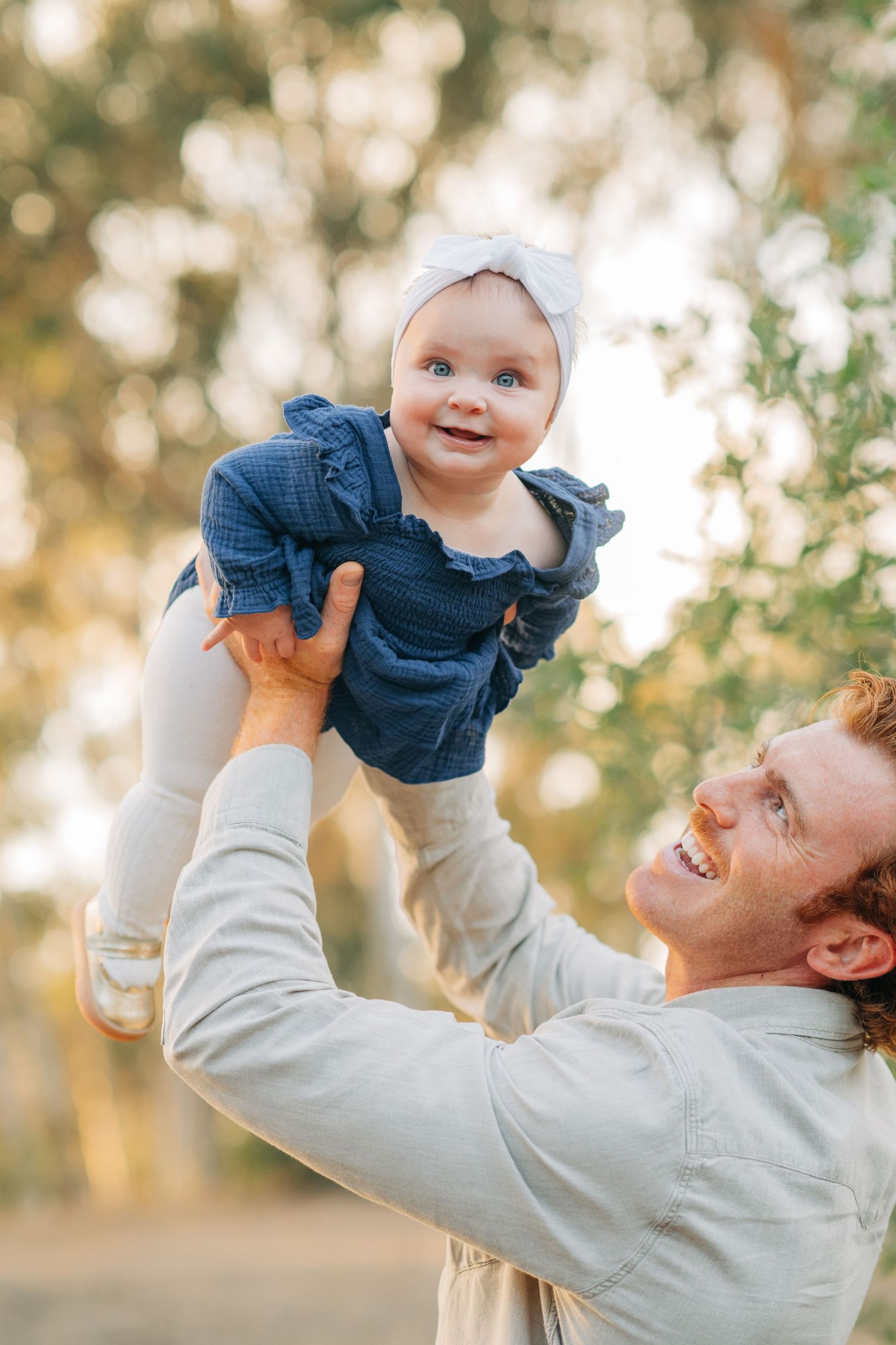 Smiling father lifting happy baby girl in blue dress and white headband outdoors in a sunny park.