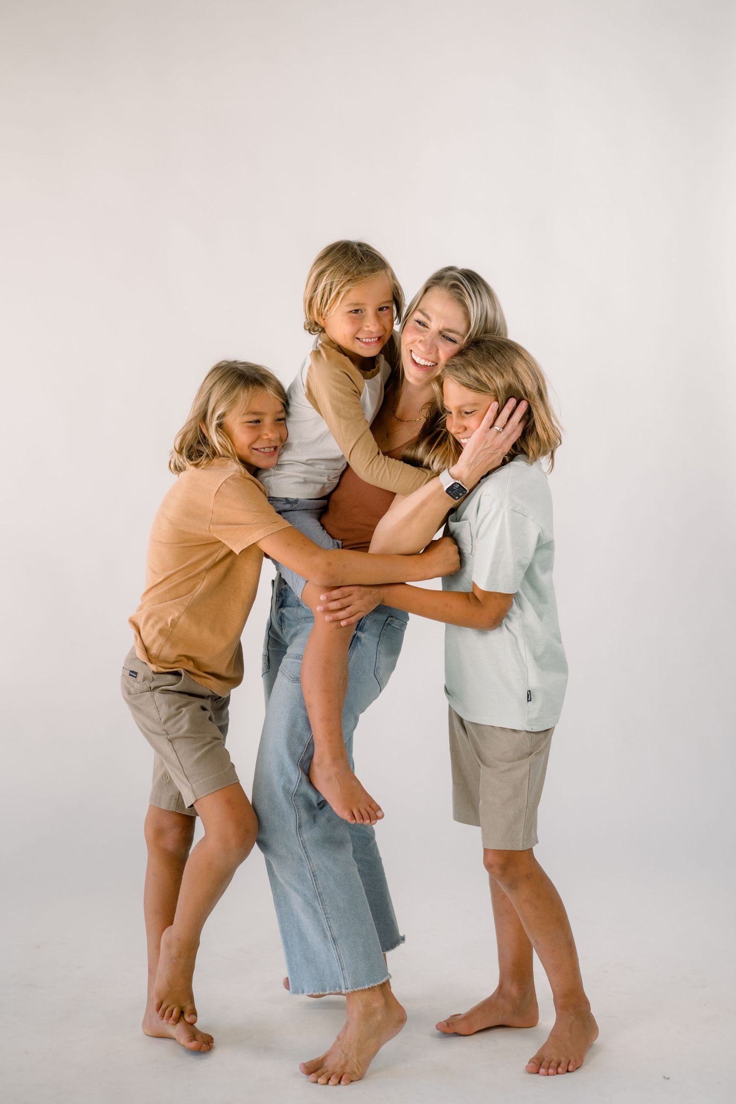 A woman and three children hug and laugh together in a bright white studio, wearing casual neutral-toned outfits.