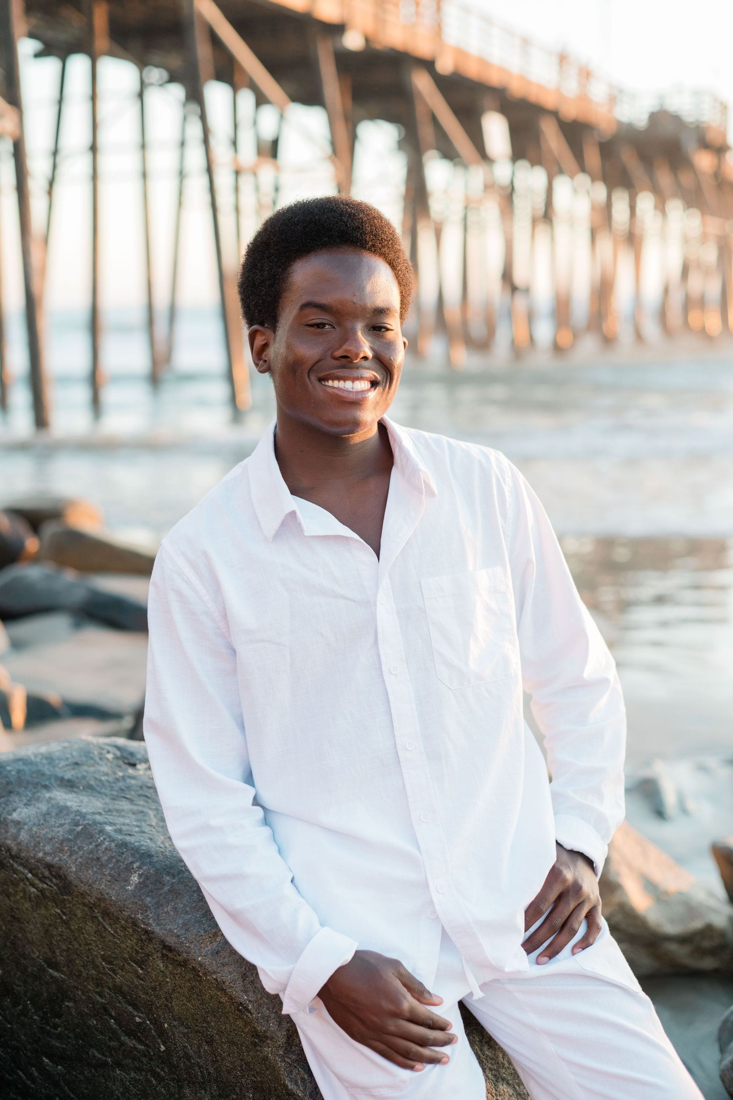 Smiling young man in white shirt and pants sitting on rocks near a beach pier at sunset.