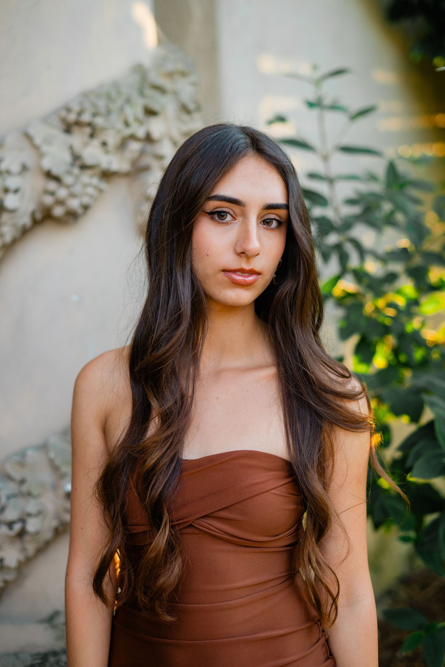 Young woman with long wavy brown hair wearing a strapless rust-colored dress, posing outdoors near greenery.