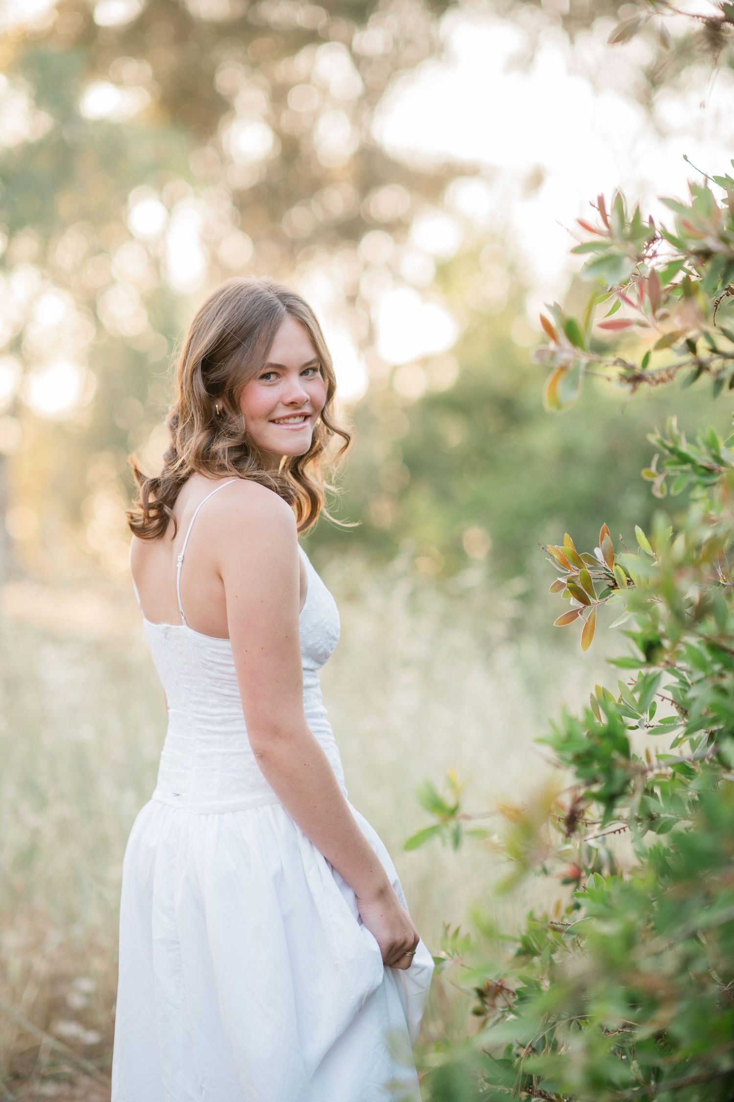 Young woman in white sundress smiling over shoulder in golden hour sunlit garden with lush green foliage.