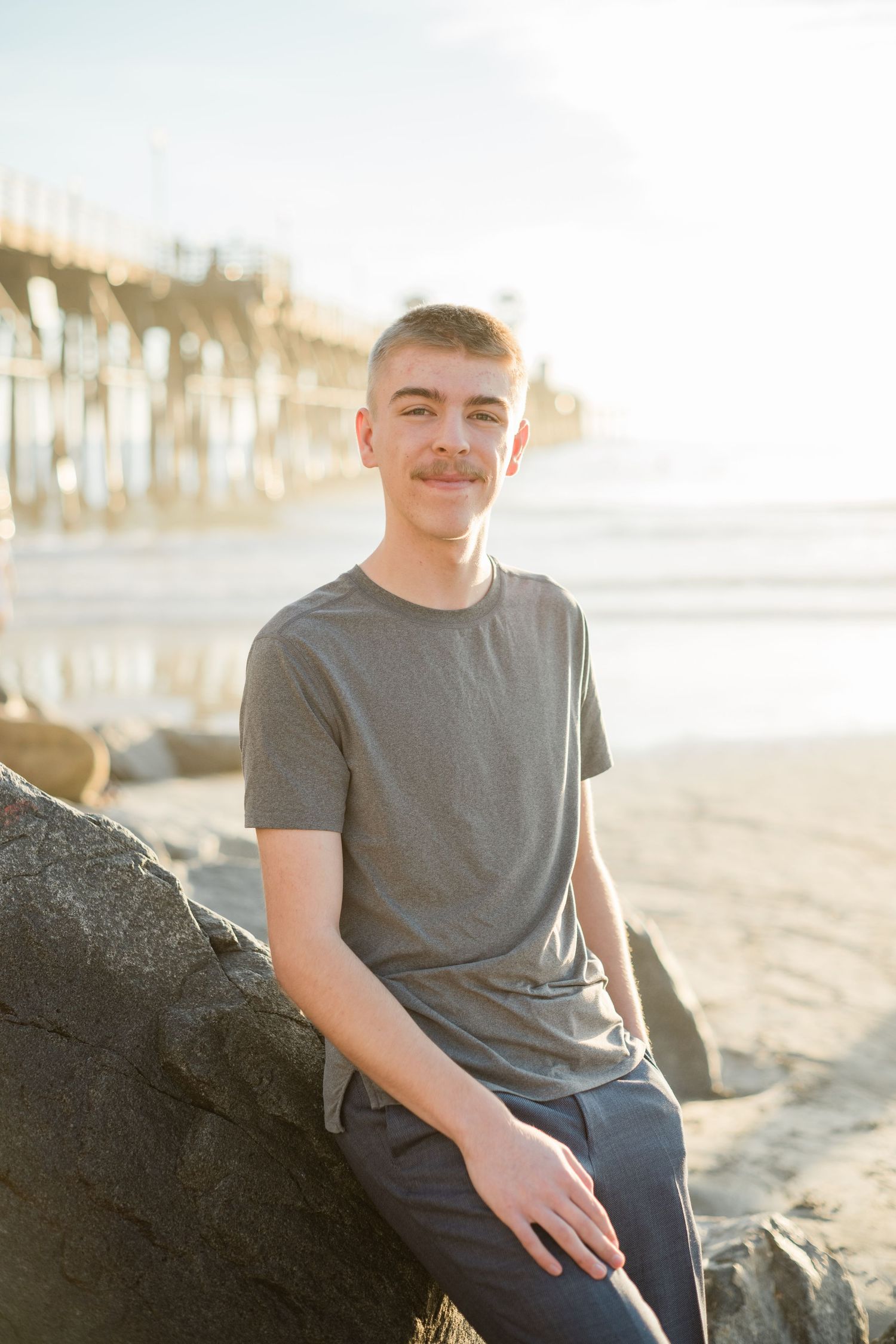 Young man with mustache sitting on rocks at sunny beach pier, wearing gray t-shirt and navy pants, smiling.