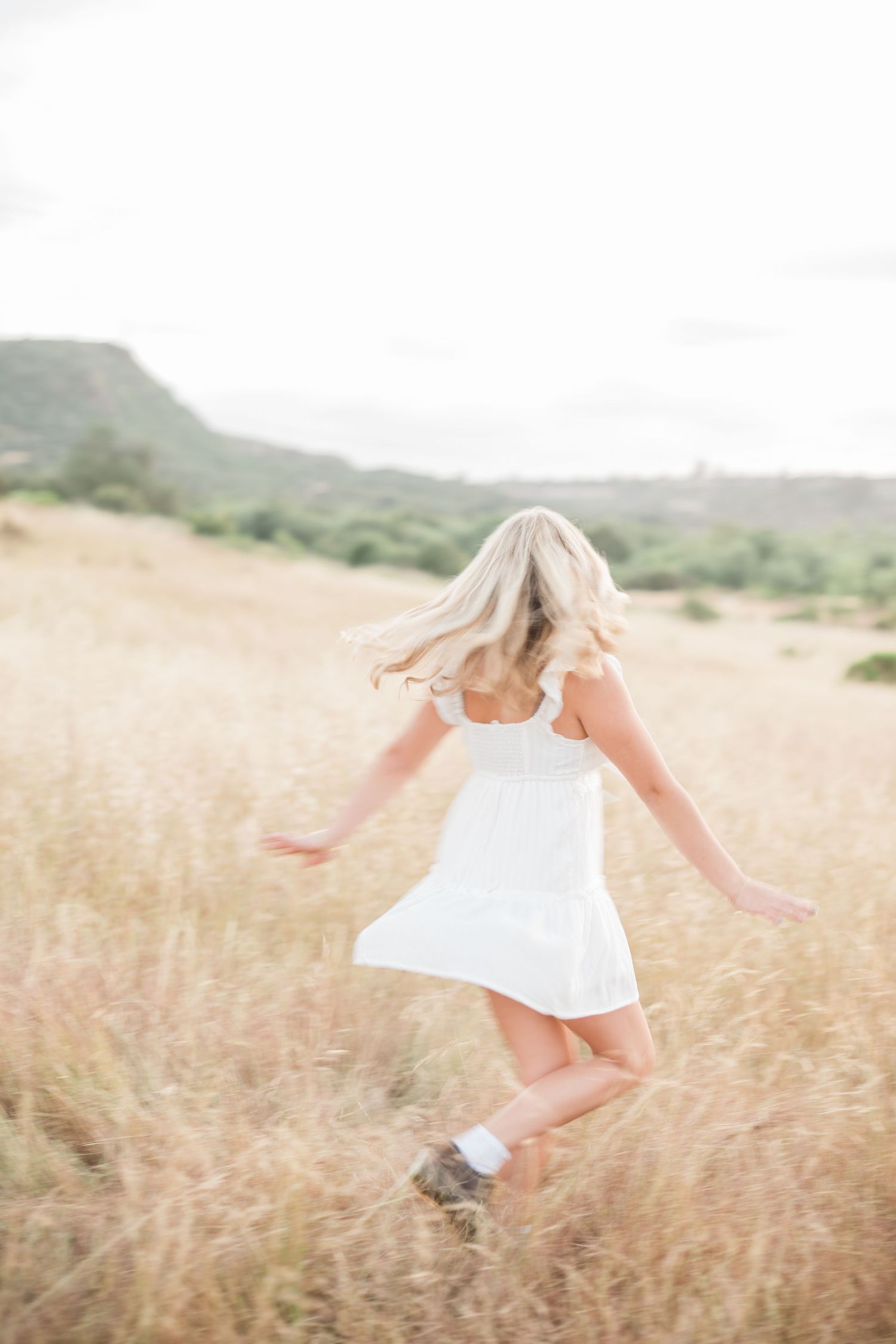 Senior session of a girl in white dress spinning joyfully through a golden dry grass field with scenic hills behind her.