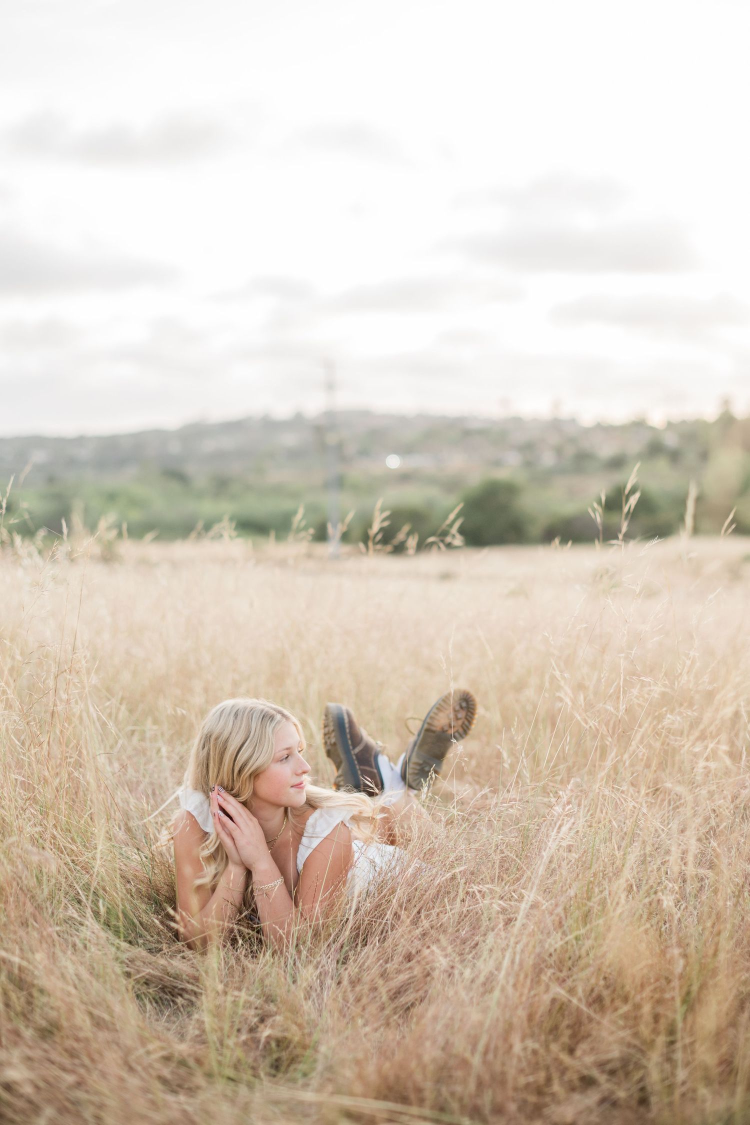 Senior sessions of a girl in white dress lying relaxed in tall golden grass field, gazing into the distance outdoors.