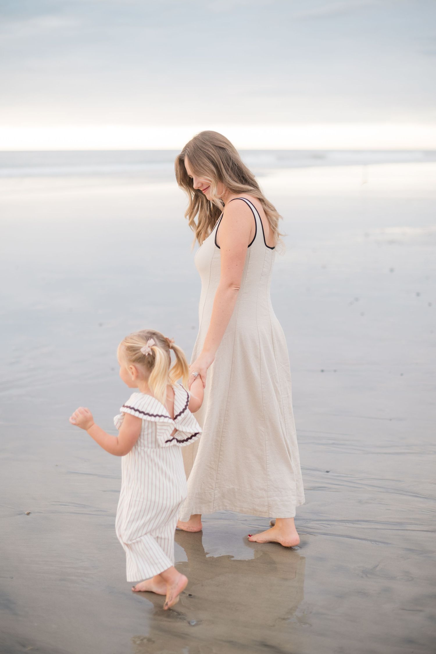 Mother and young daughter in matching outfits walking hand-in-hand barefoot on a calm beach at dusk.