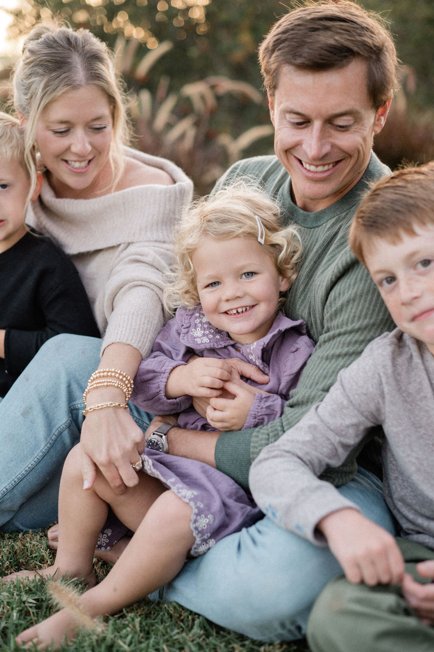 Happy family of five sitting together on grass smiling in warm outdoor setting.