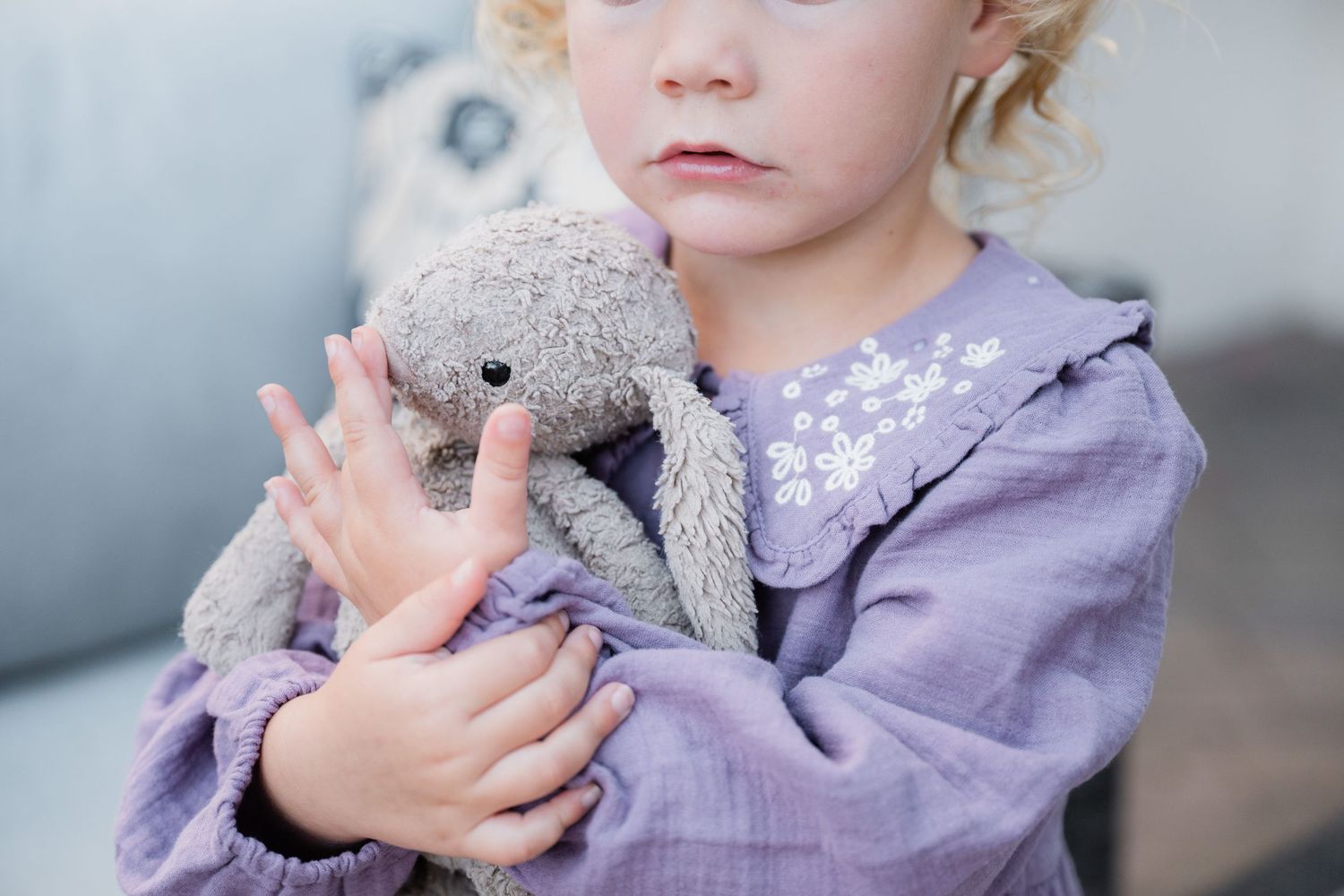 Young girl in purple dress hugging a gray stuffed bunny toy, with a soft blurred background.