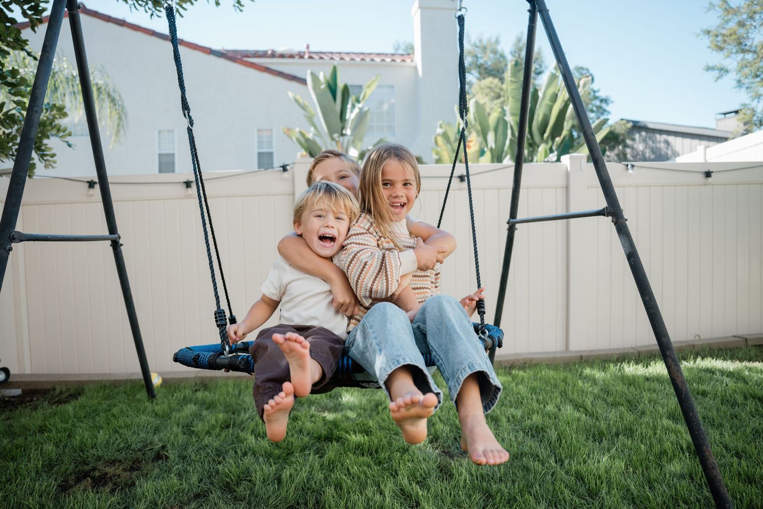 Two happy children sitting together on a backyard swing set on a sunny day, smiling and laughing outdoors.