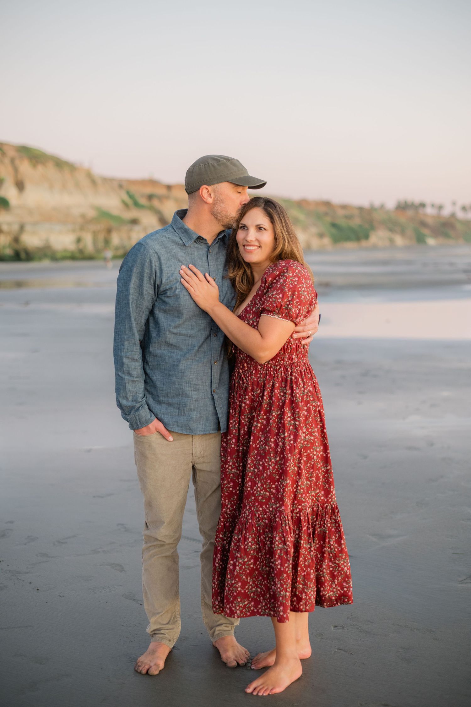 Couple embracing on a sandy beach at sunset, man in blue shirt and cap kissing woman in red floral dress.