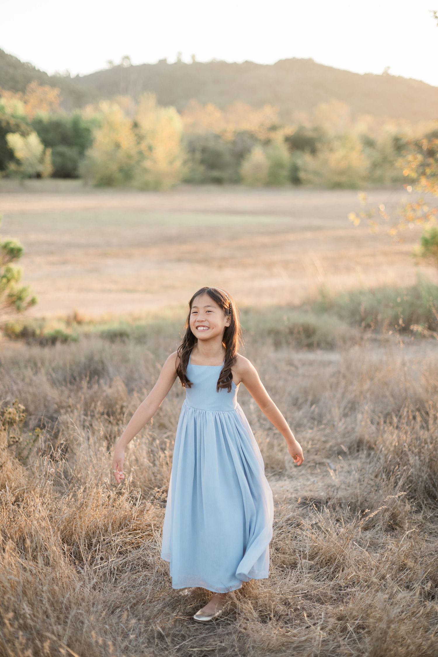 A smiling young Asian girl in a light blue dress stands in a golden field with hills in the background.