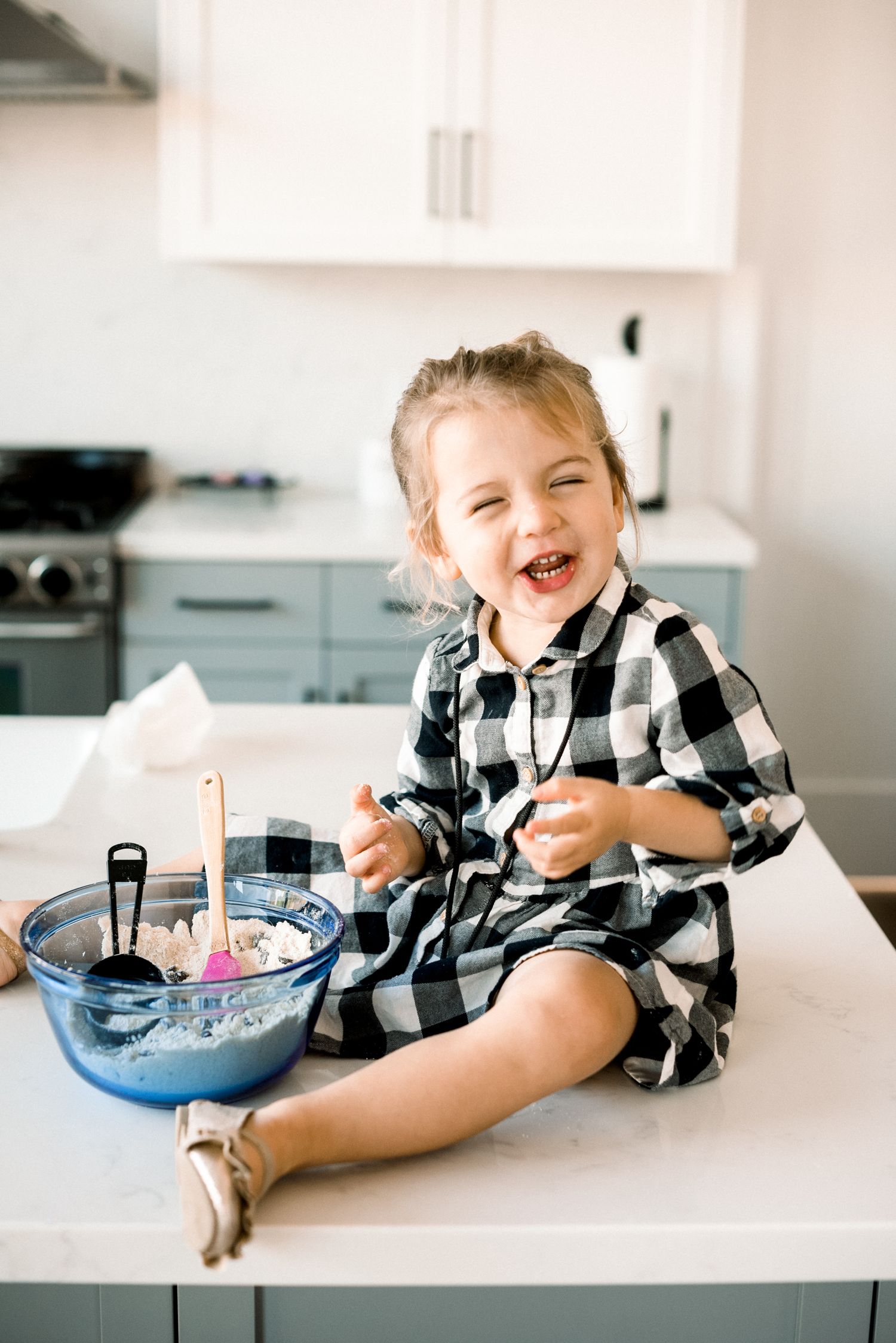 Happy toddler girl in plaid dress sitting on kitchen counter laughing while helping bake with mixing bowl.