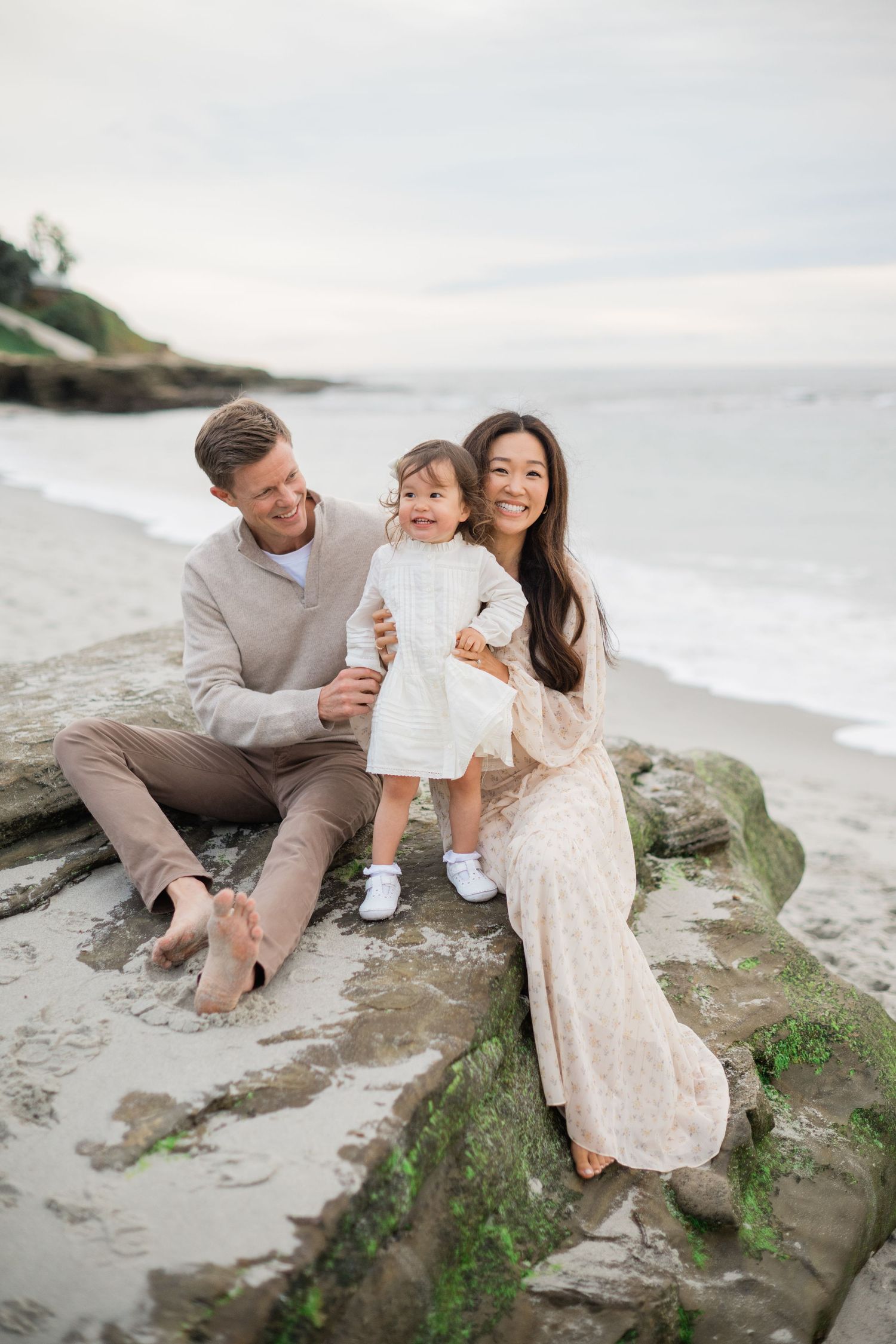 Happy family sitting on beach rocks, parents with smiling toddler in white dress, ocean view behind them.