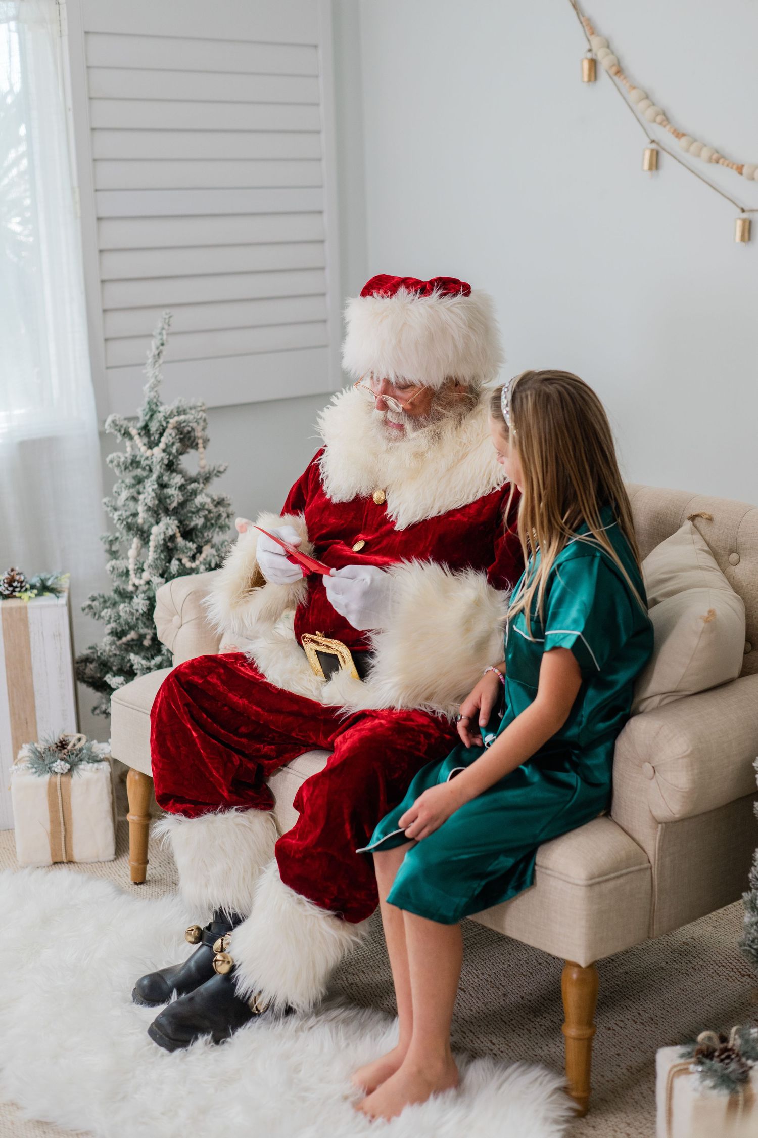 Girl in green dress sits with Santa Claus on beige sofa near Christmas tree with gifts.