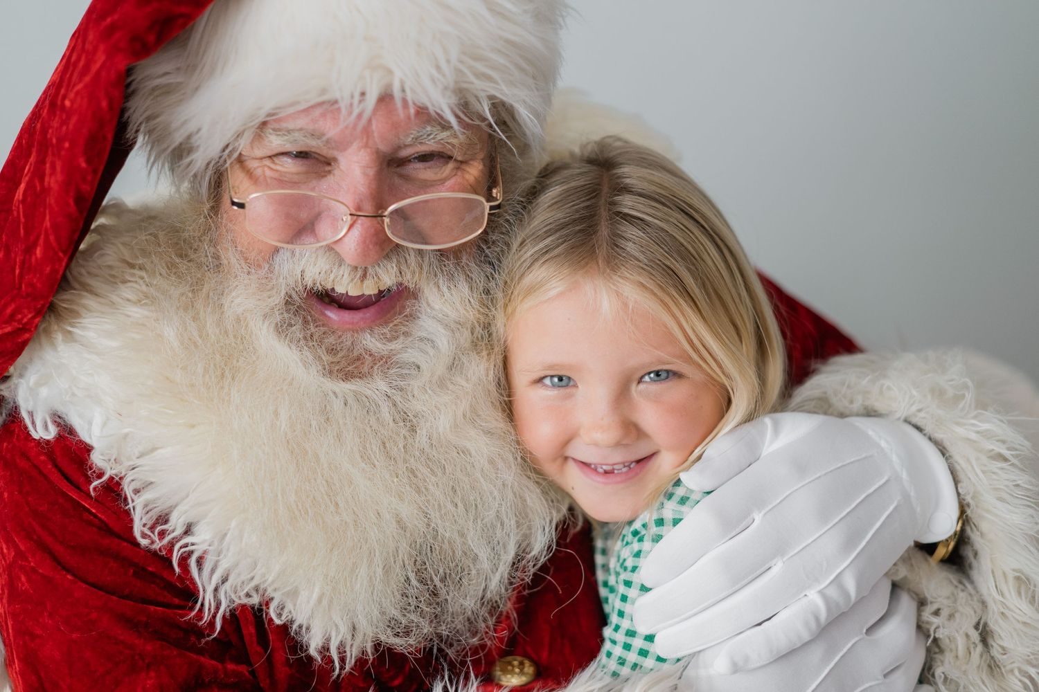 A jolly Santa Claus in red suit hugs a smiling young blonde girl with blue eyes during a festive Christmas photo.