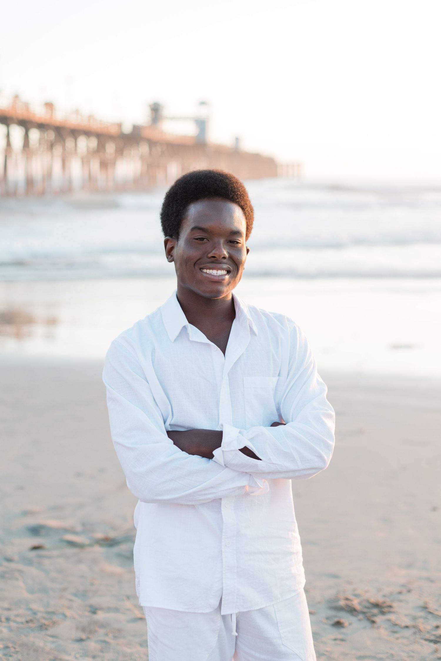 Smiling young man in white outfit standing on beach with arms crossed, pier visible in background.
