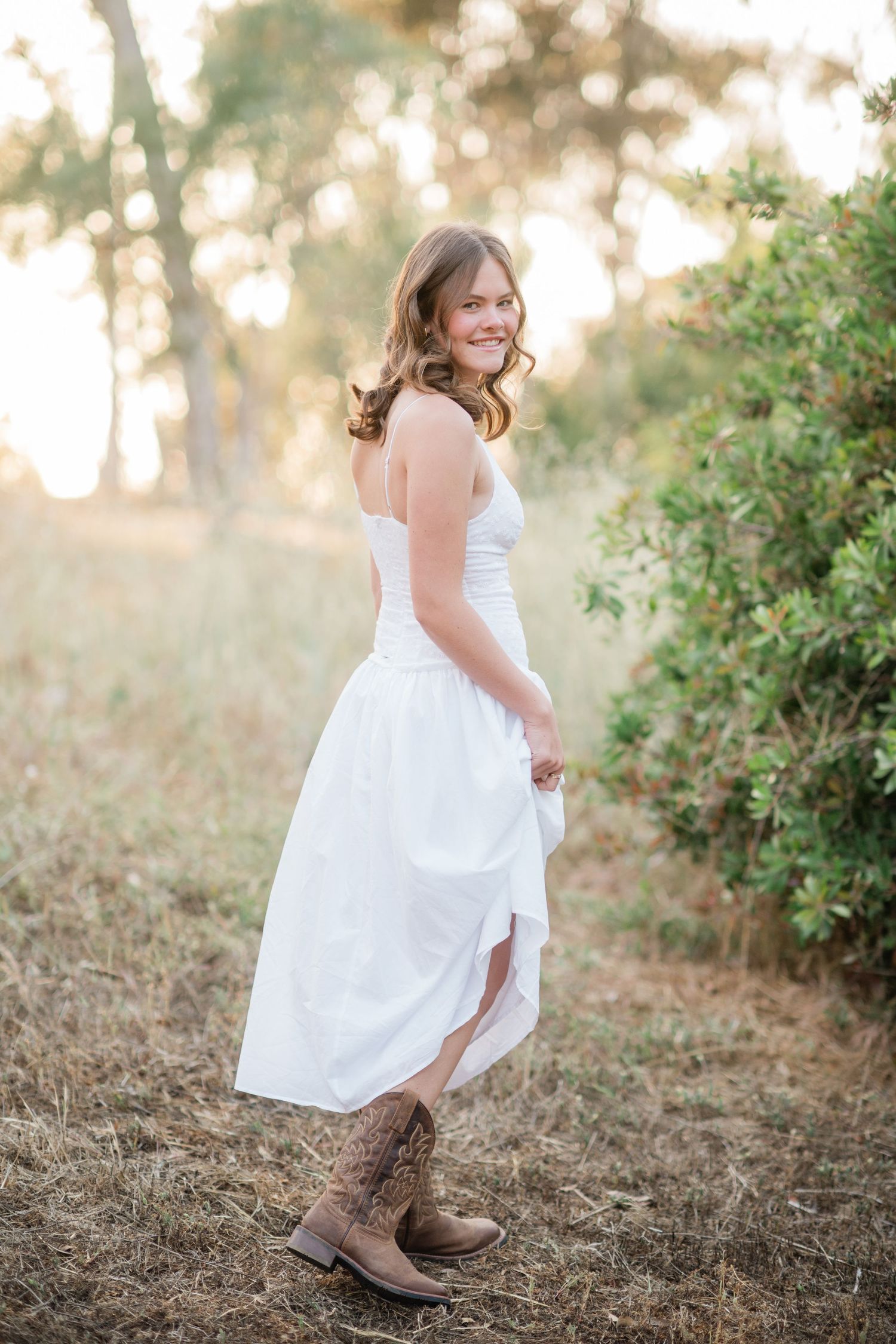 Young woman in white strapless dress and cowboy boots smiling outdoors in golden hour light.