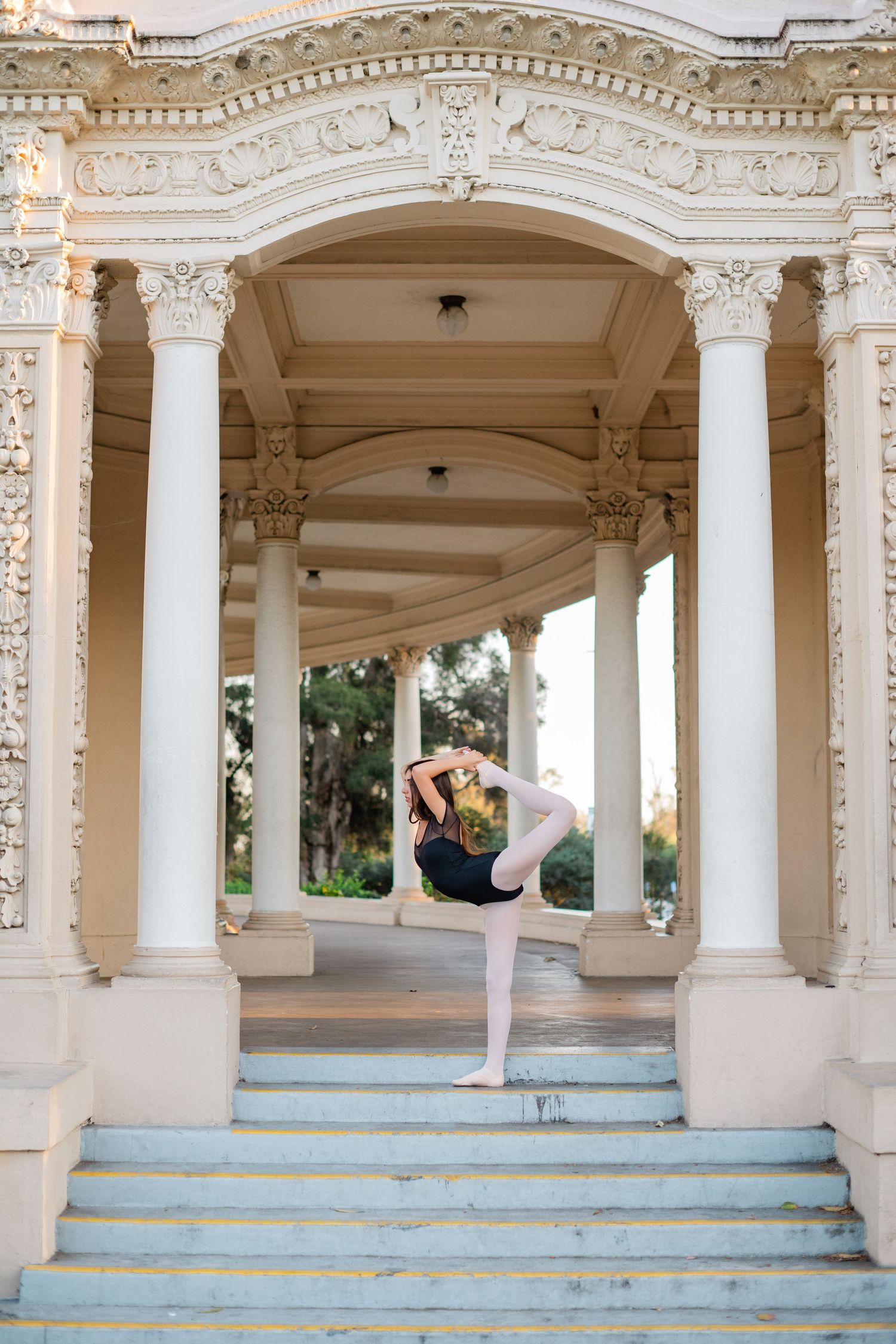 Dancer poses gracefully in Lord of the Dance yoga pose on steps of ornate white classical colonnade building.