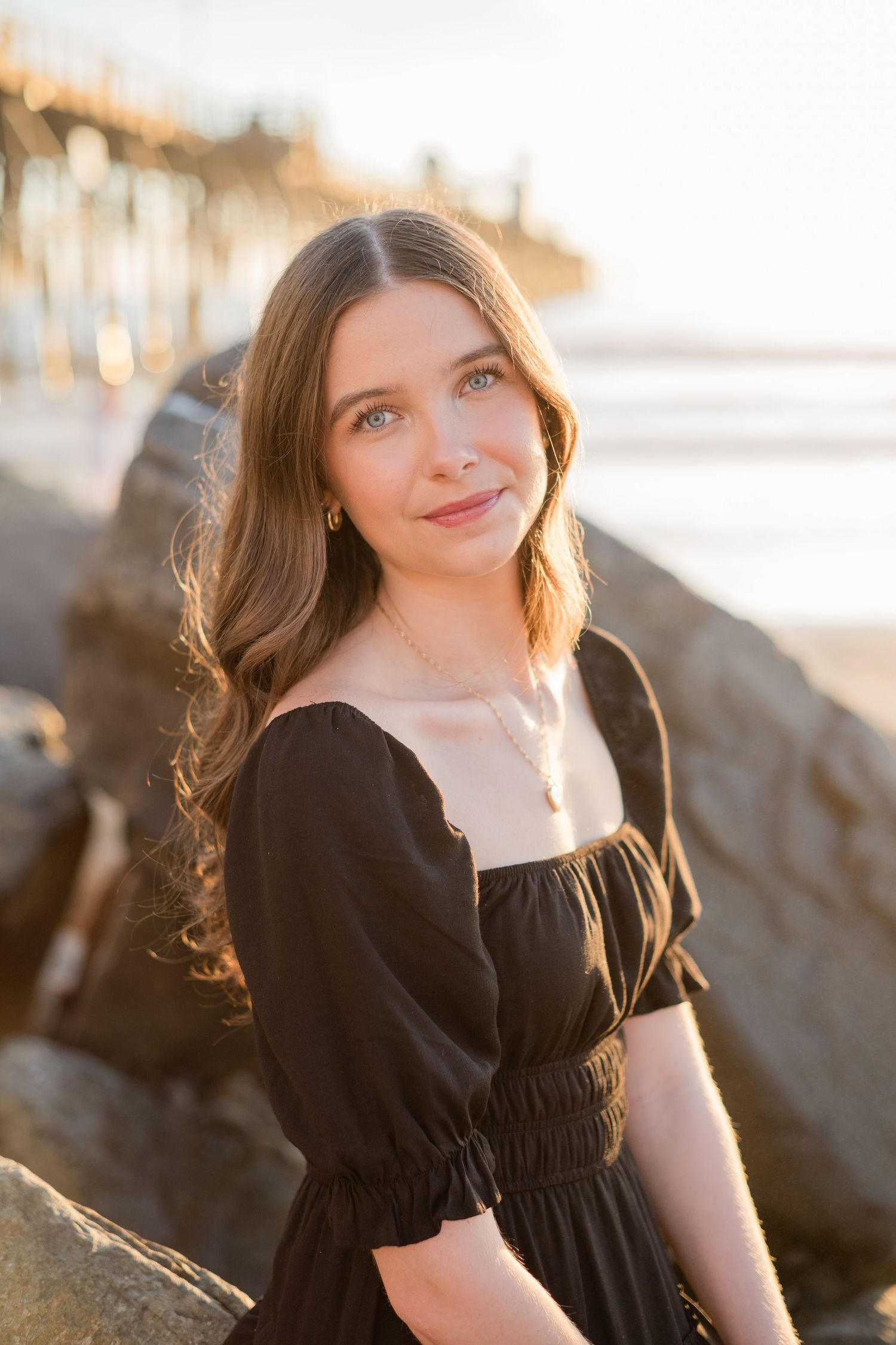 Young woman in black dress poses on beach rocks at golden hour with pier in background.