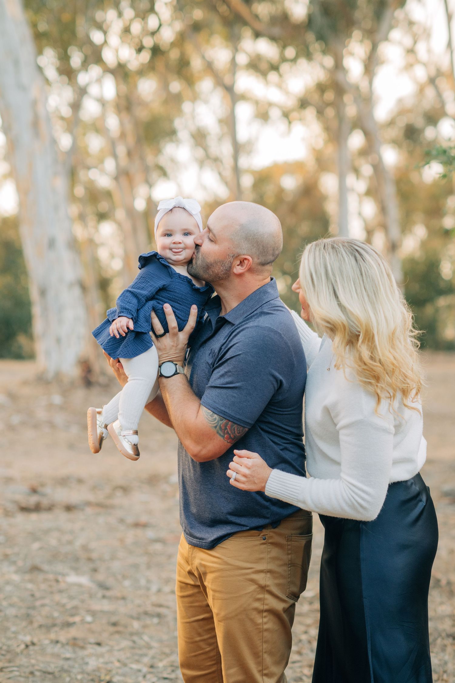 A bald father kisses his smiling toddler while blonde mother embraces them in a sunlit outdoor park setting.