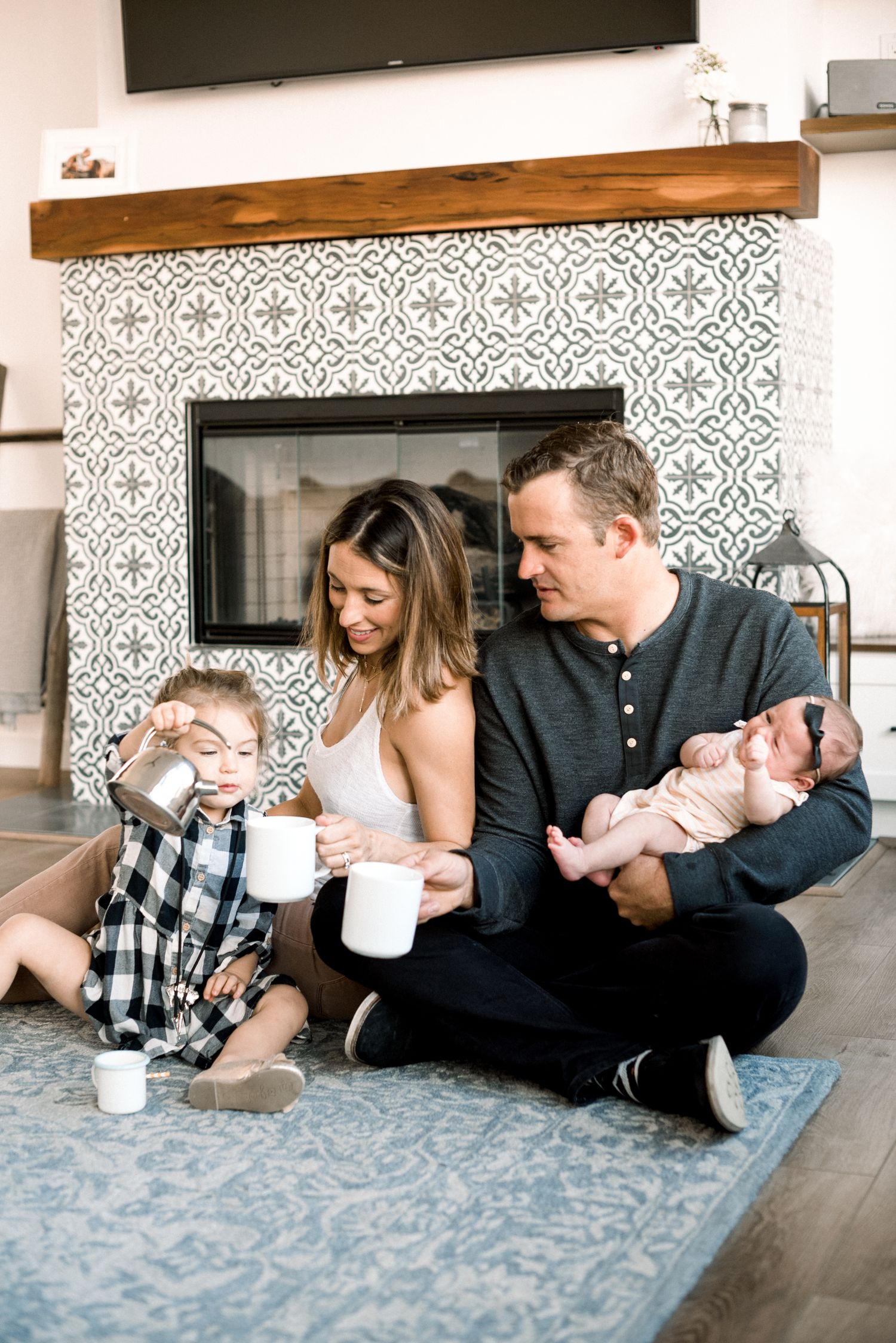 Family of four sitting by fireplace, parents holding mugs while dad cradles newborn and toddler plays.