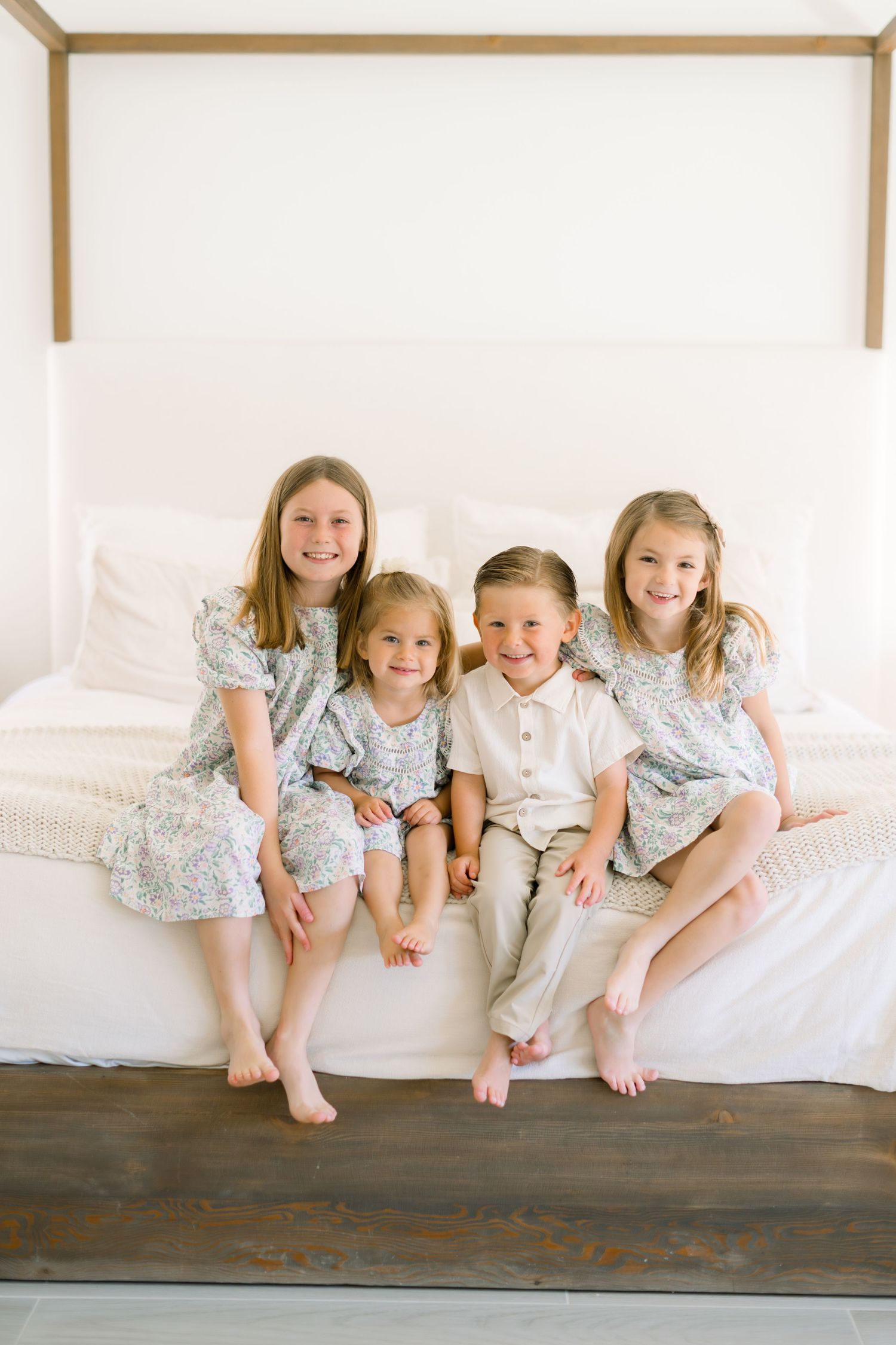 Four young children sitting together on a white bed, smiling, dressed in soft floral and neutral outfits.