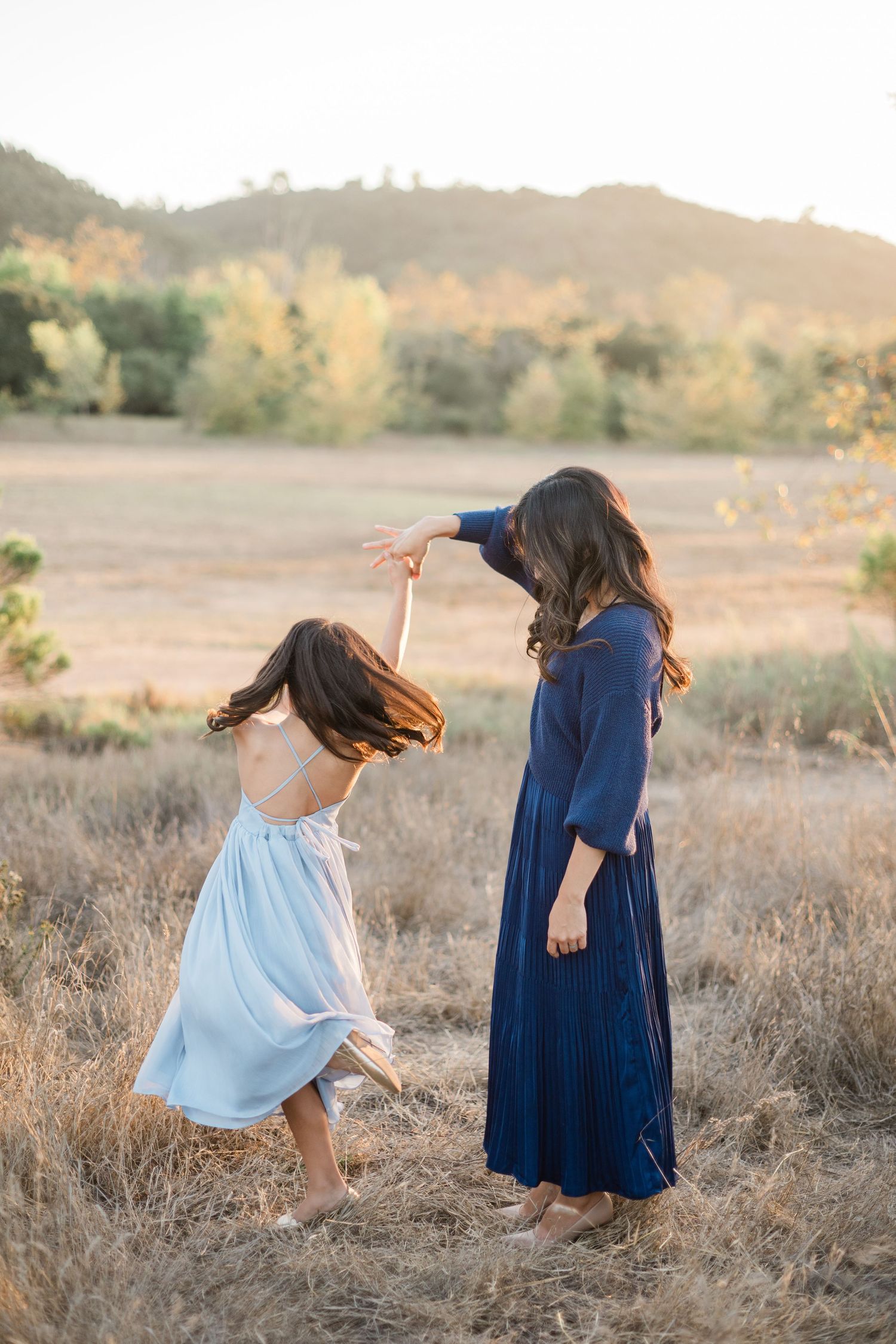 A mother twirls her young daughter in a sunlit field, both wearing flowing blue dresses at golden hour.