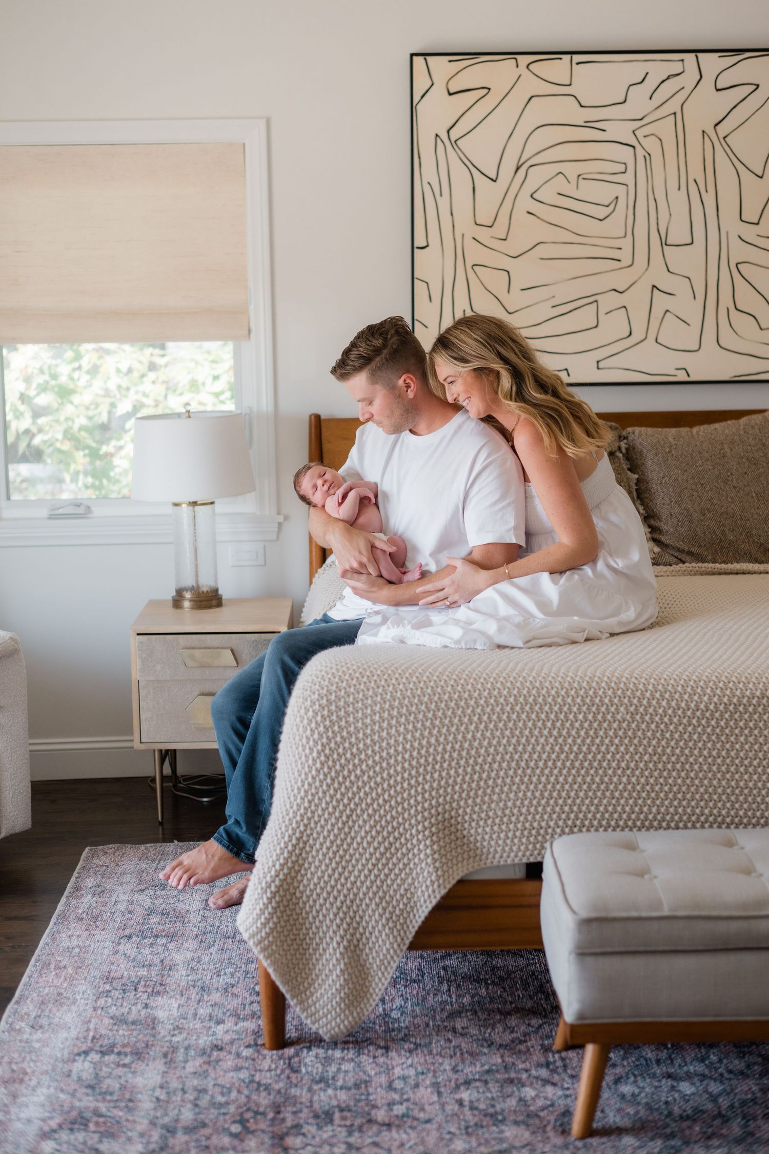 Young couple sitting on bed with newborn baby in bright modern bedroom with abstract wall art.