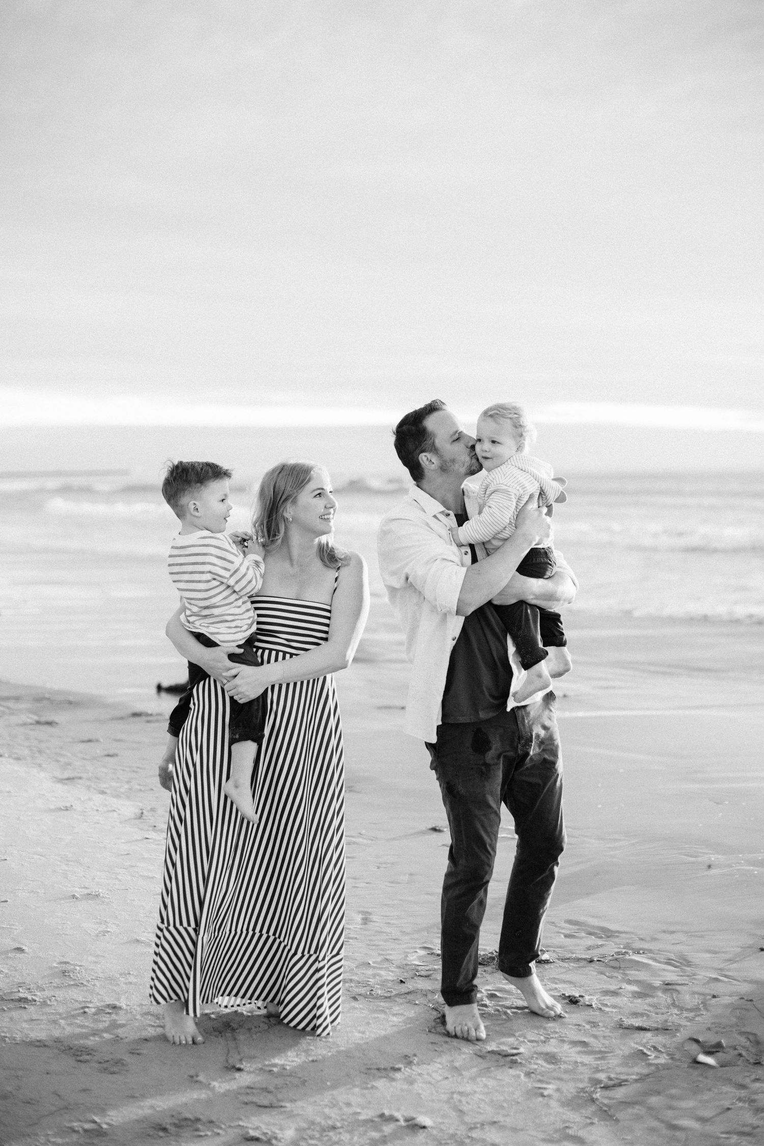Black and white photo of a happy family of four on a beach, parents holding young children, waves in background.